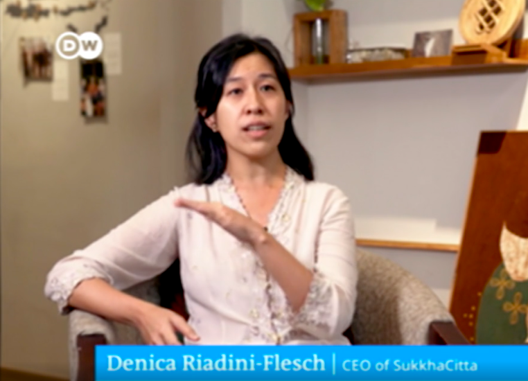 Denica Riadini-Flesch speaking in an interview, labeled as "CEO of SukkhaCitta." The setting includes a chair and wall decor.