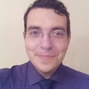 Close-up photo of a man with glasses, short dark hair, wearing a purple shirt and black tie, smiling lightly, against a light background.