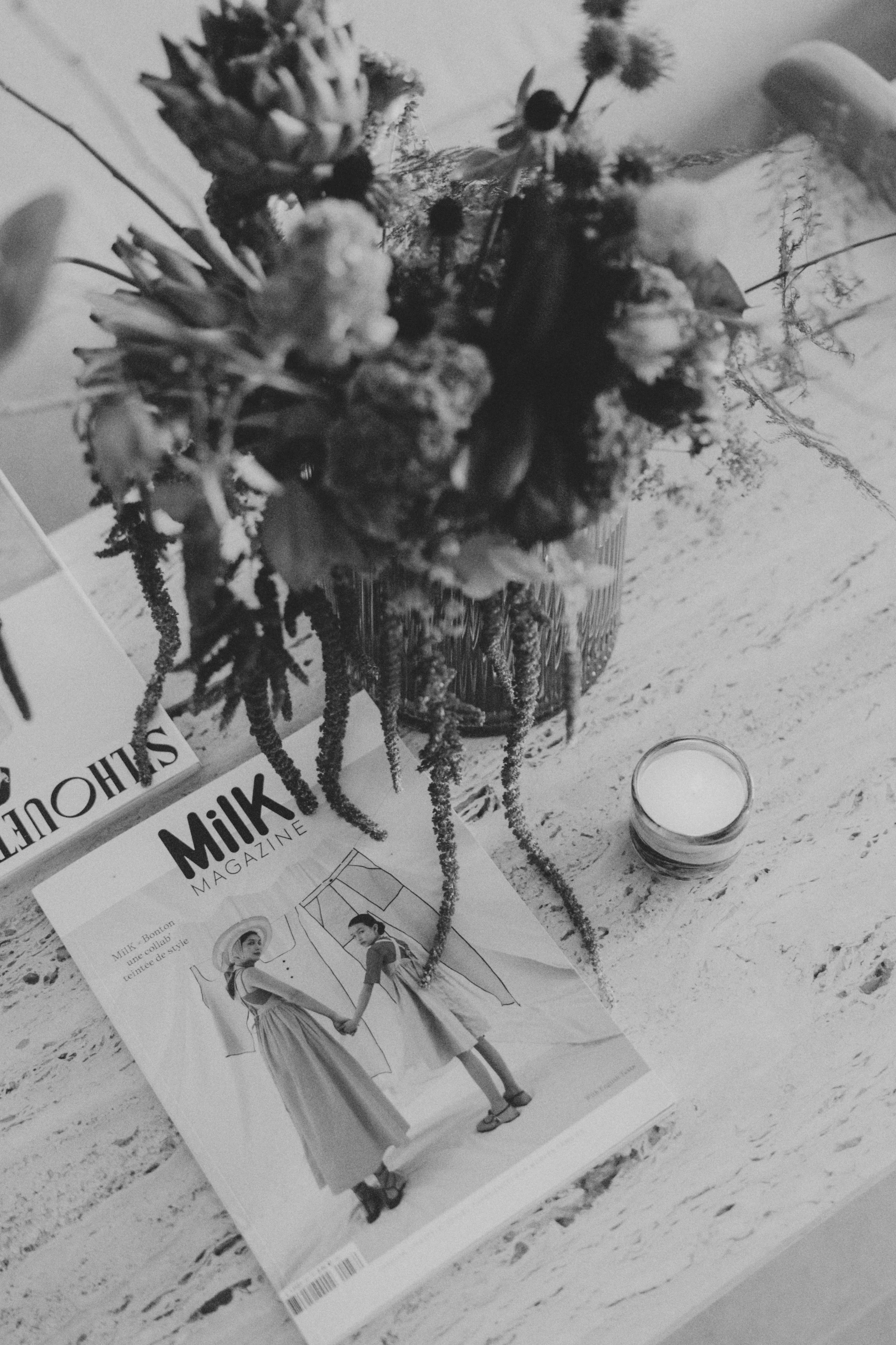 Un bouquet de fleurs dans un vase placé sur une table, avec un magazine intitulé 'Milk Magazine' et une bougie à côté.