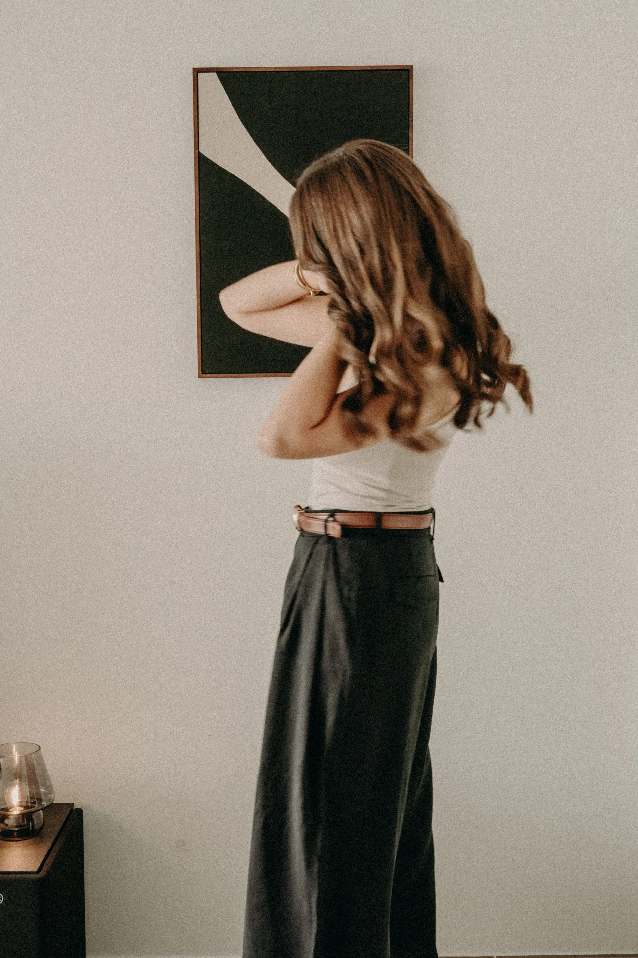 Une femme aux cheveux bruns bouclés se tient devant un tableau noir abstrait sur un mur blanc, portant un haut blanc et une longue jupe noire.