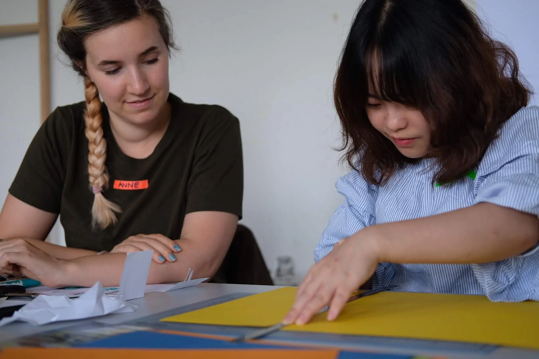 Two women working together at a table, one with a braid and a name tag that reads 'Anne,' and the other with dark hair, focusing on colorful papers on the table.