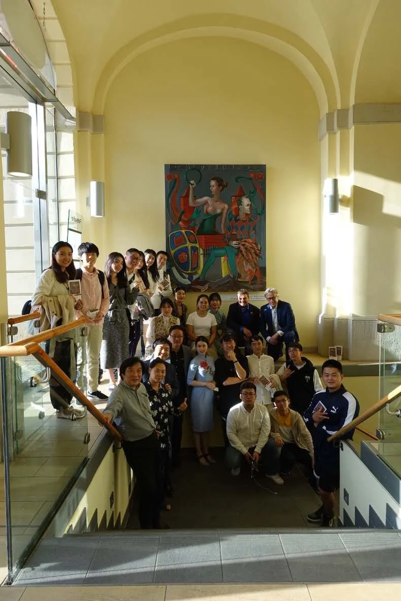 Group of people standing on stairs in a yellow-walled indoor space with a large colorful painting of a woman holding a diamond and a mask hanging on the wall behind them.