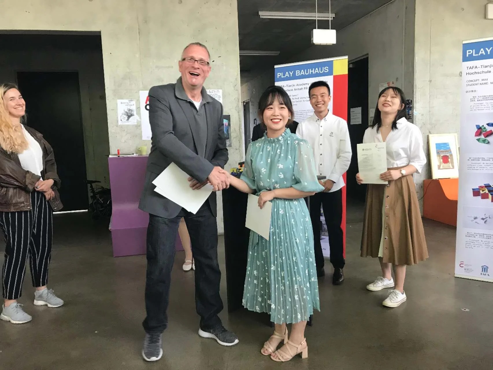 A group of people at an event, with a man in a gray blazer shaking hands with a woman in a light blue dress, all smiling.