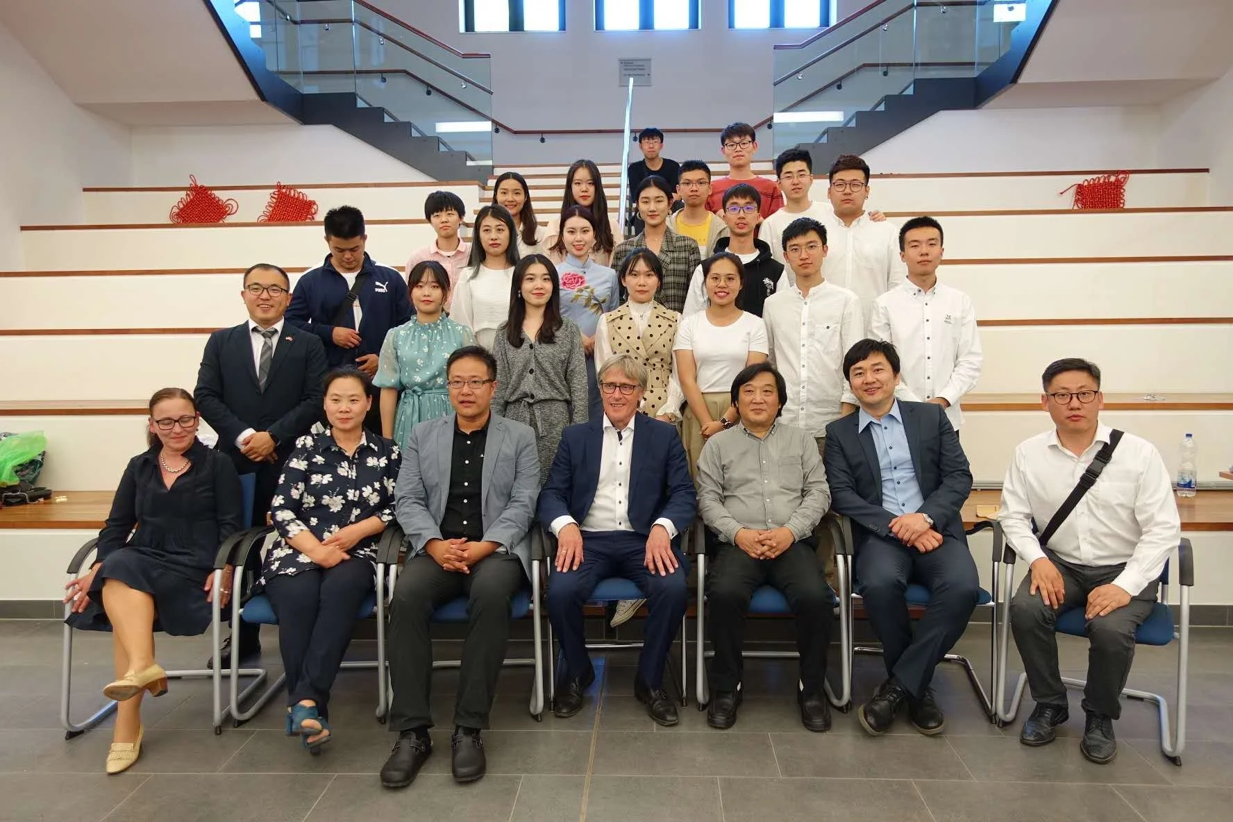 Group photo of 21 people, including students and faculty, posing in a lecture hall with stairs and railings in the background.