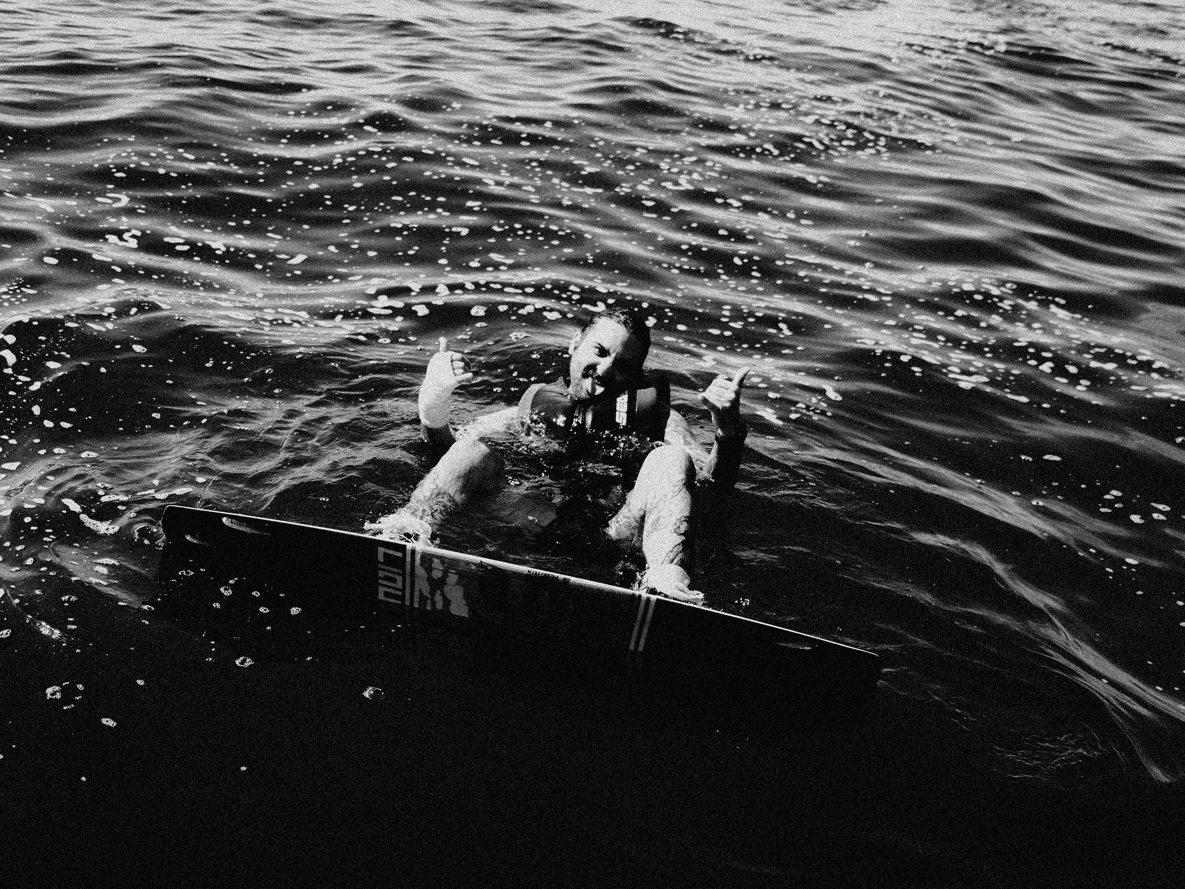 Person smiling in water holding a skateboard and giving a thumbs-up while making a tongue out gesture.