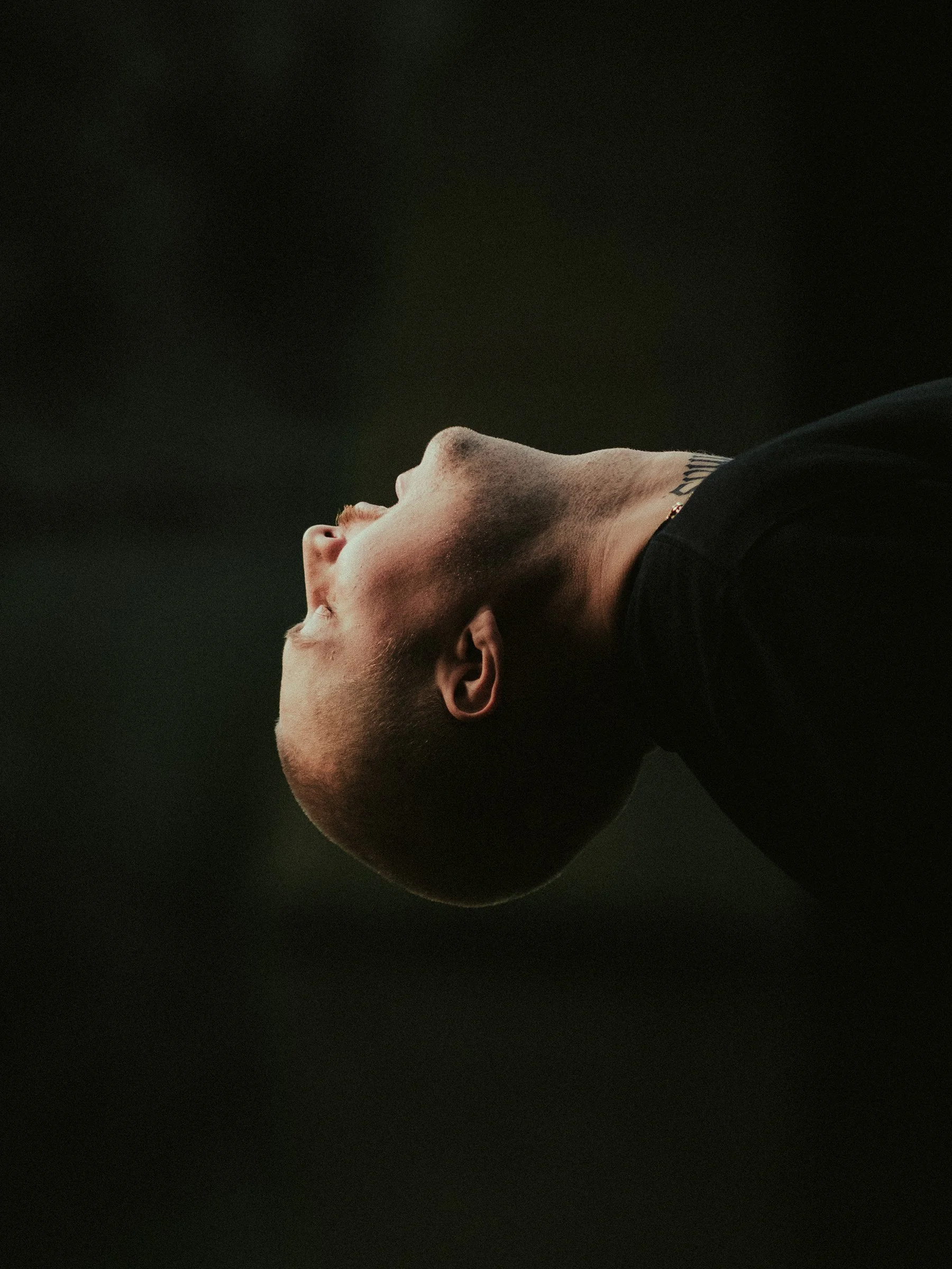 A close-up profile of a man with a shaved head, looking upward with a dark, blurred background.
