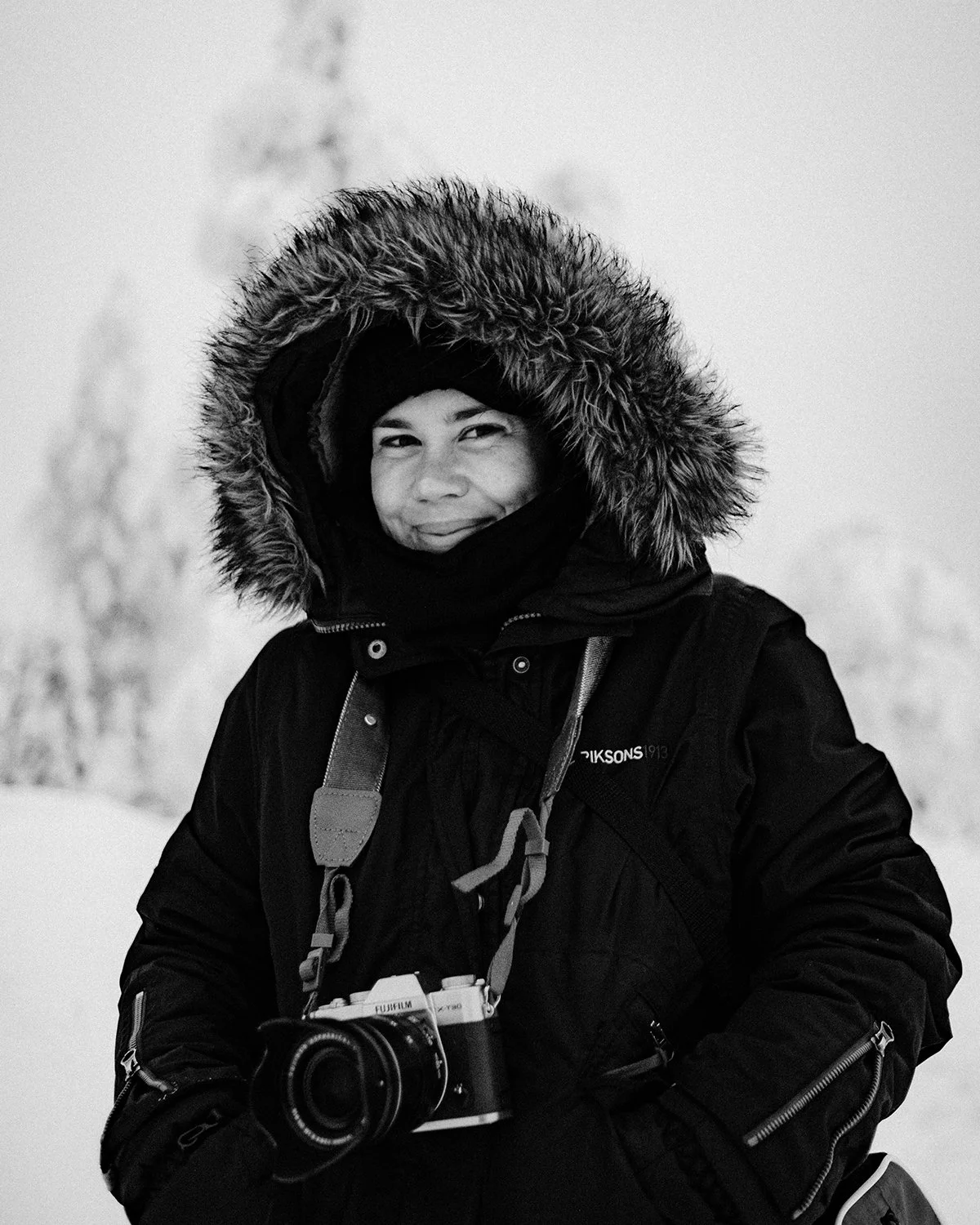 Woman in winter clothing and fur-lined hood holding a camera in a snowy landscape.