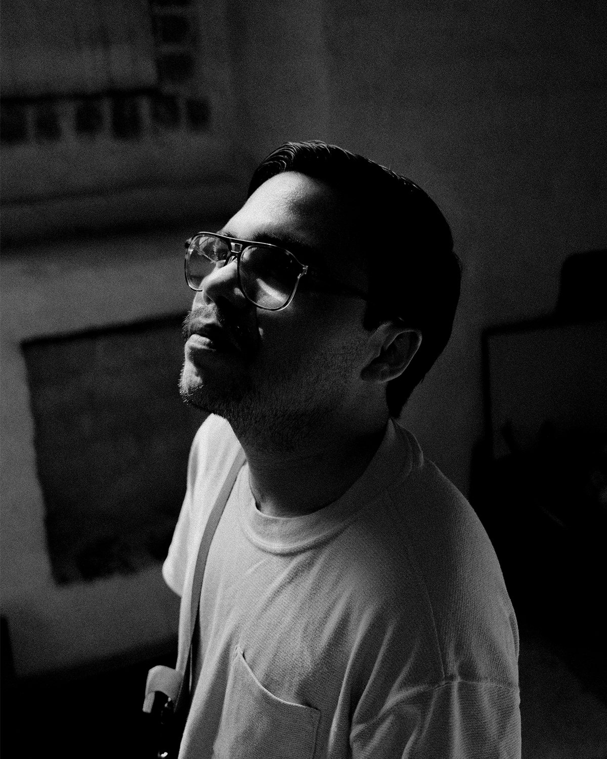 Black and white photo of a man with glasses, short hair, and facial hair, looking slightly upwards, in a dimly lit room.