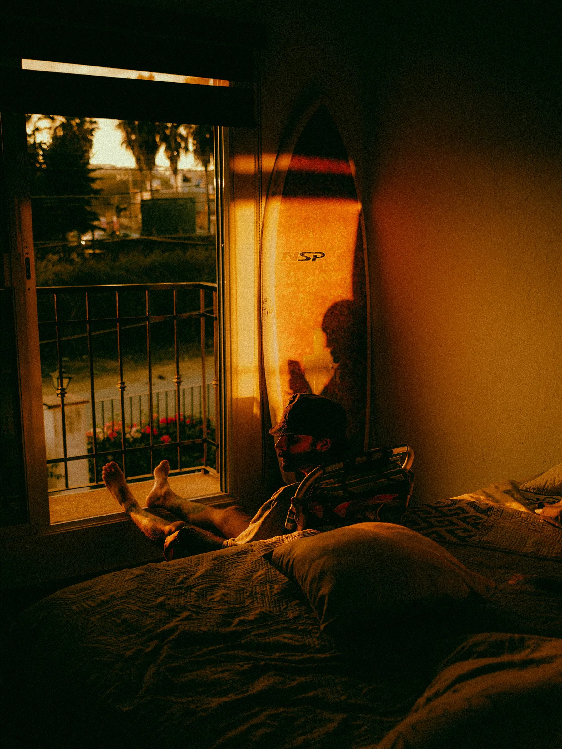 A person sitting by a window with warm sunlight shining on them, a surfboard leaning against the wall, and a balcony with flowers outside.