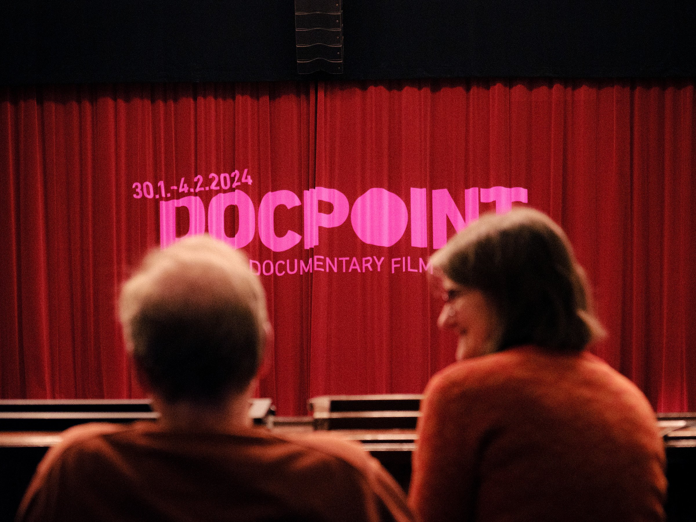 Audience members sitting in front of a red curtain with a screen displaying the text 'DOCPOINT Documentary Film' and dates '30.1-4.2.2024' at a film festival.