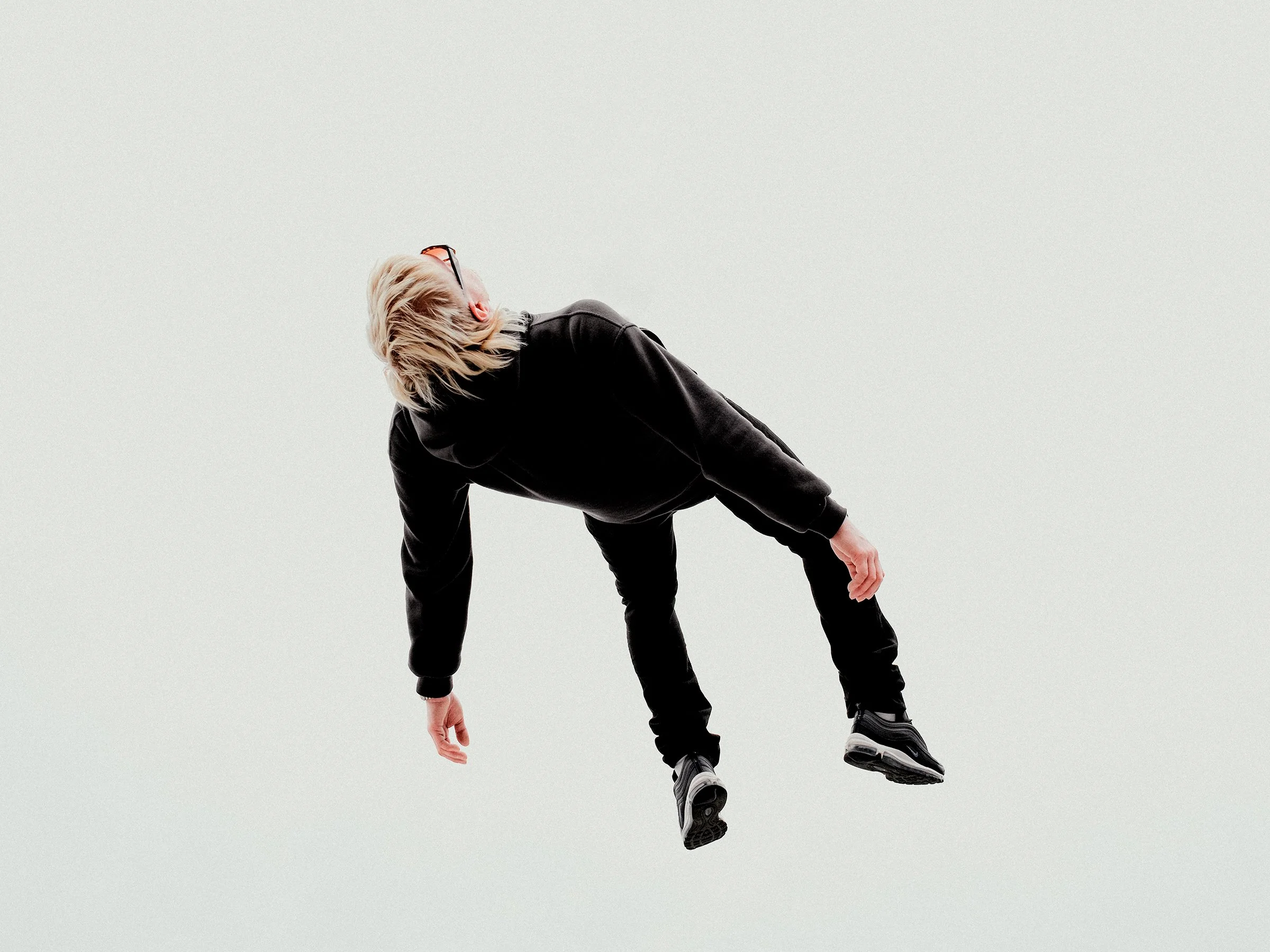 Person with blonde hair wearing glasses and black clothing, leaning backward as if falling or floating against a plain, light background.