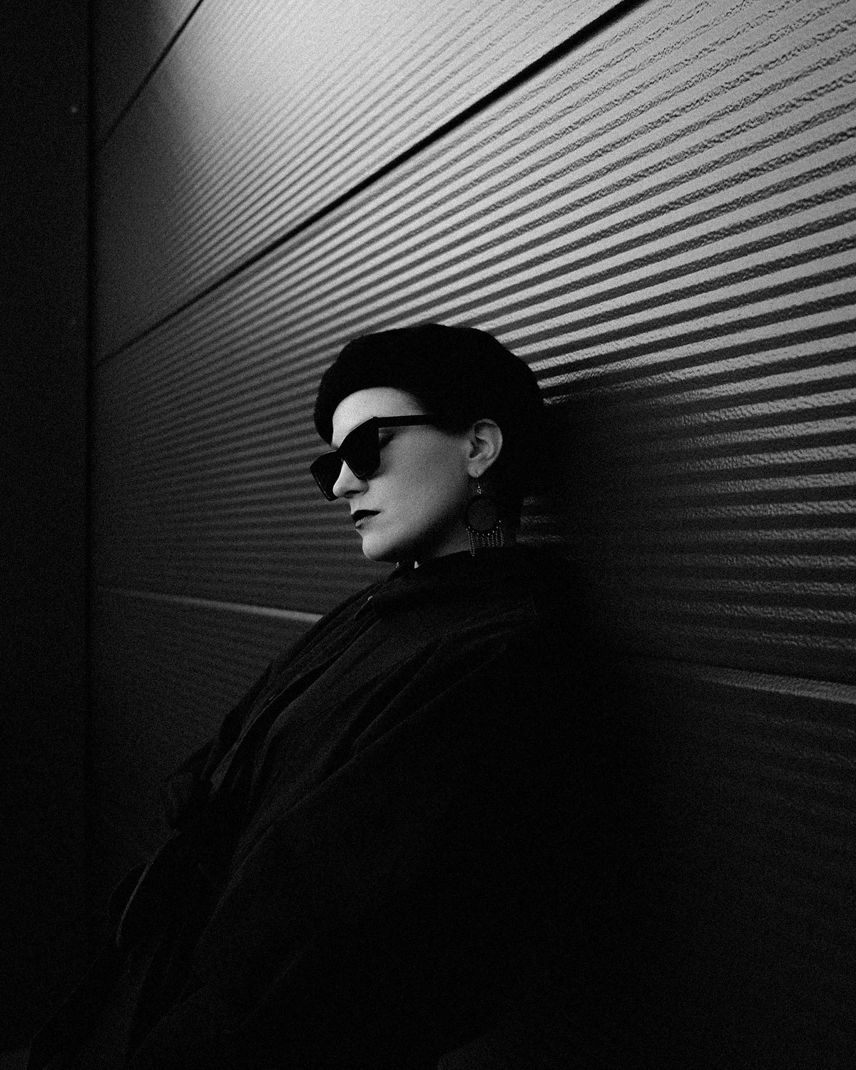 Black and white photo of a woman with dark hair, glasses, and earrings, leaning against a textured wall, with her eyes closed and hair blowing to the side.