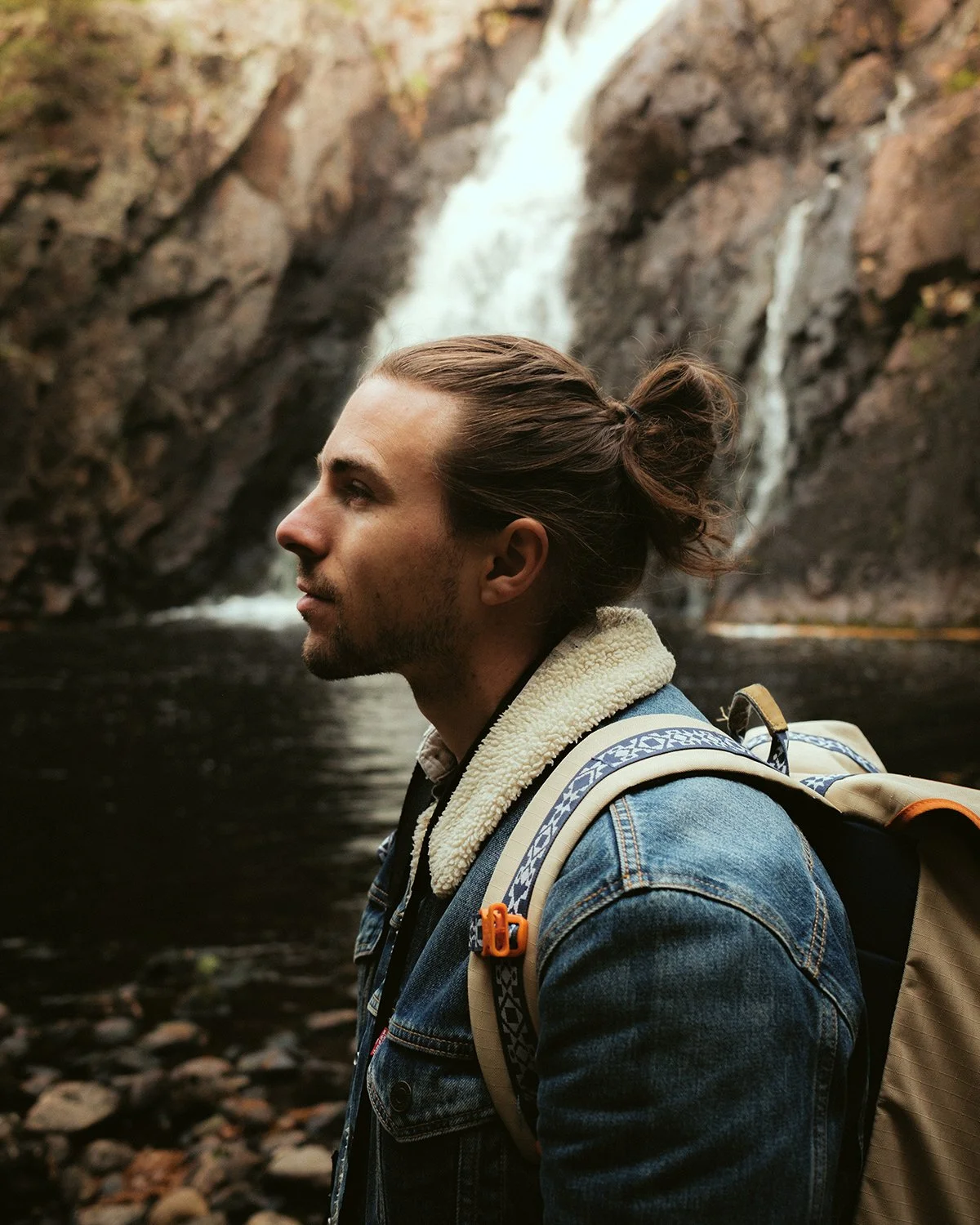 A man with long hair tied back, wearing a denim jacket and backpack, stands near a river with a waterfall in the background.