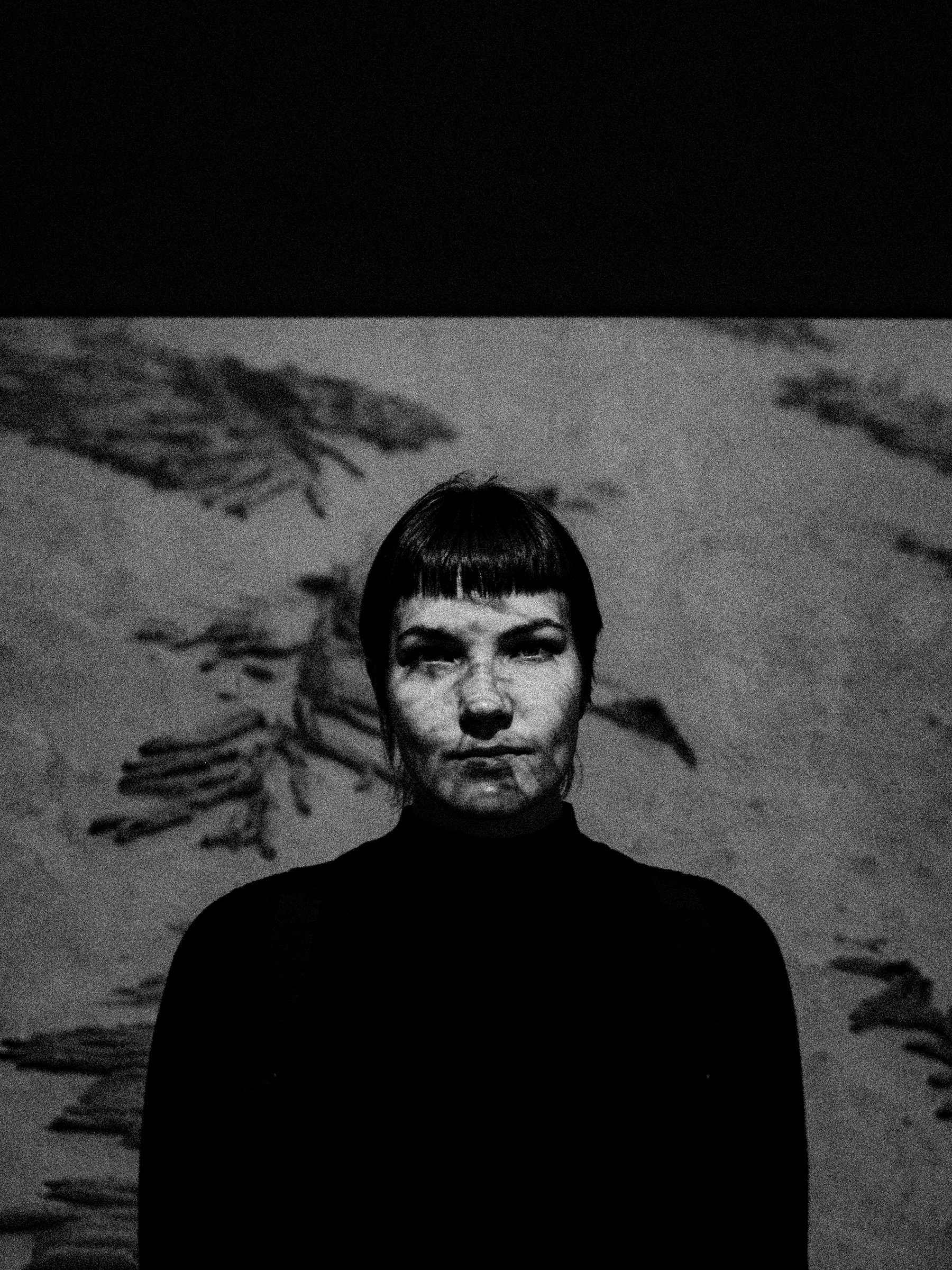 A woman with short, dark hair and a serious expression stands in front of a wall with shadows of plants cast behind her. The photo is in black and white.