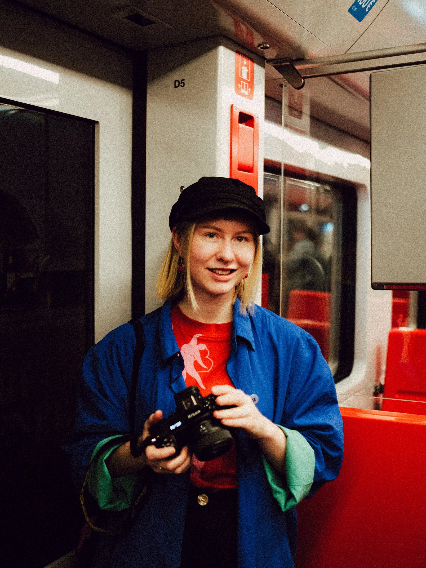 A young woman with blonde hair, wearing a black cap, blue jacket, and red shirt, holding a camera inside a train.