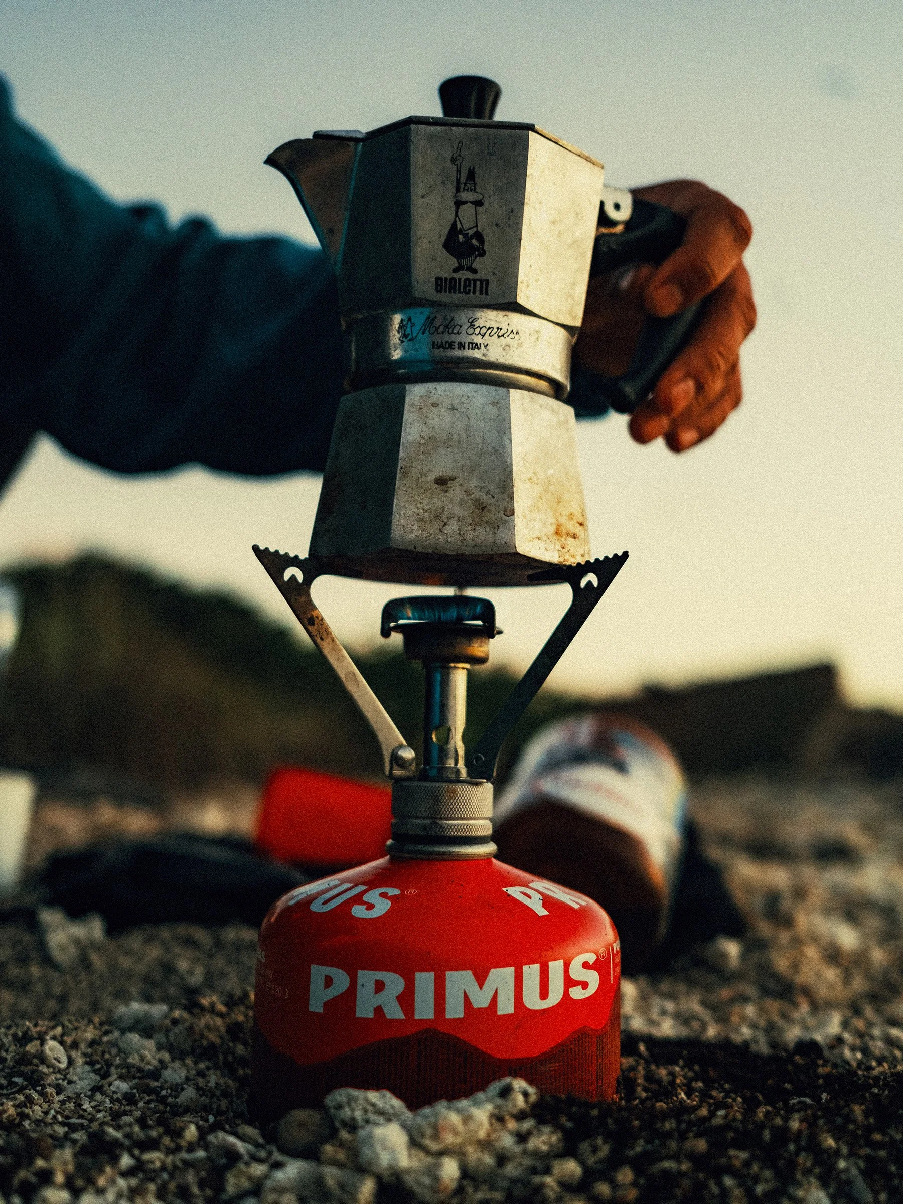 A portable camping stove with a red fuel canister on rocky ground, with a hand lighting it near a coffee maker.