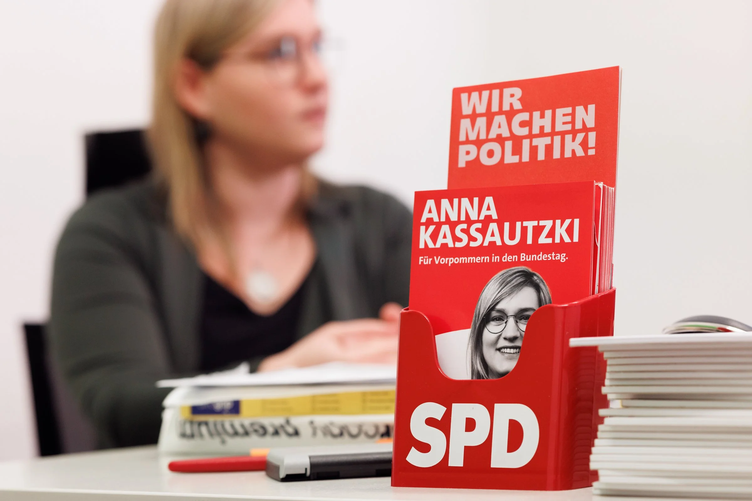 Blurred image of a woman sitting at a table with a red political brochure in the foreground that says "WIR MACHEN POLITIK!" and features a black-and-white photo of a smiling woman with glasses. The woman at the table appears to be reading or looking 
