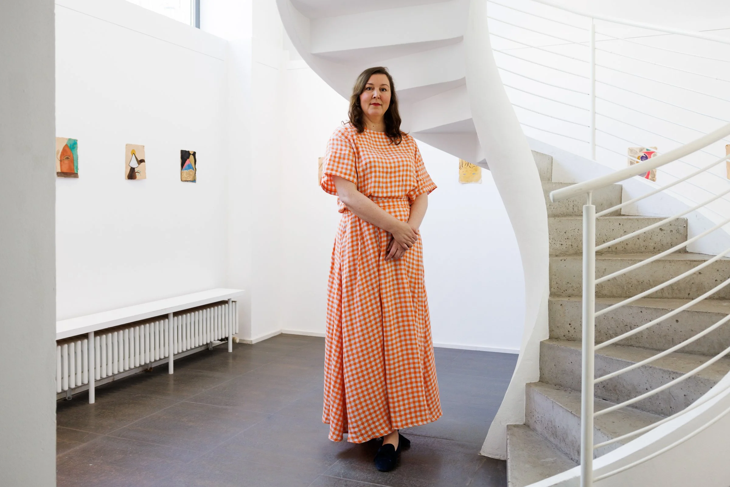 A woman in a long orange gingham dress stands near a staircase in an art gallery. The gallery has white walls, a radiator, and artwork hanging on the walls.