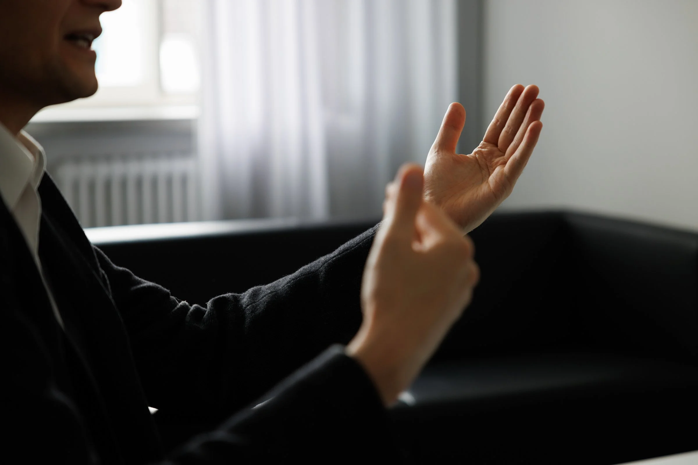 A person with dark skin is smiling and gesturing with their hand, possibly in conversation, with a window and curtains in the background.