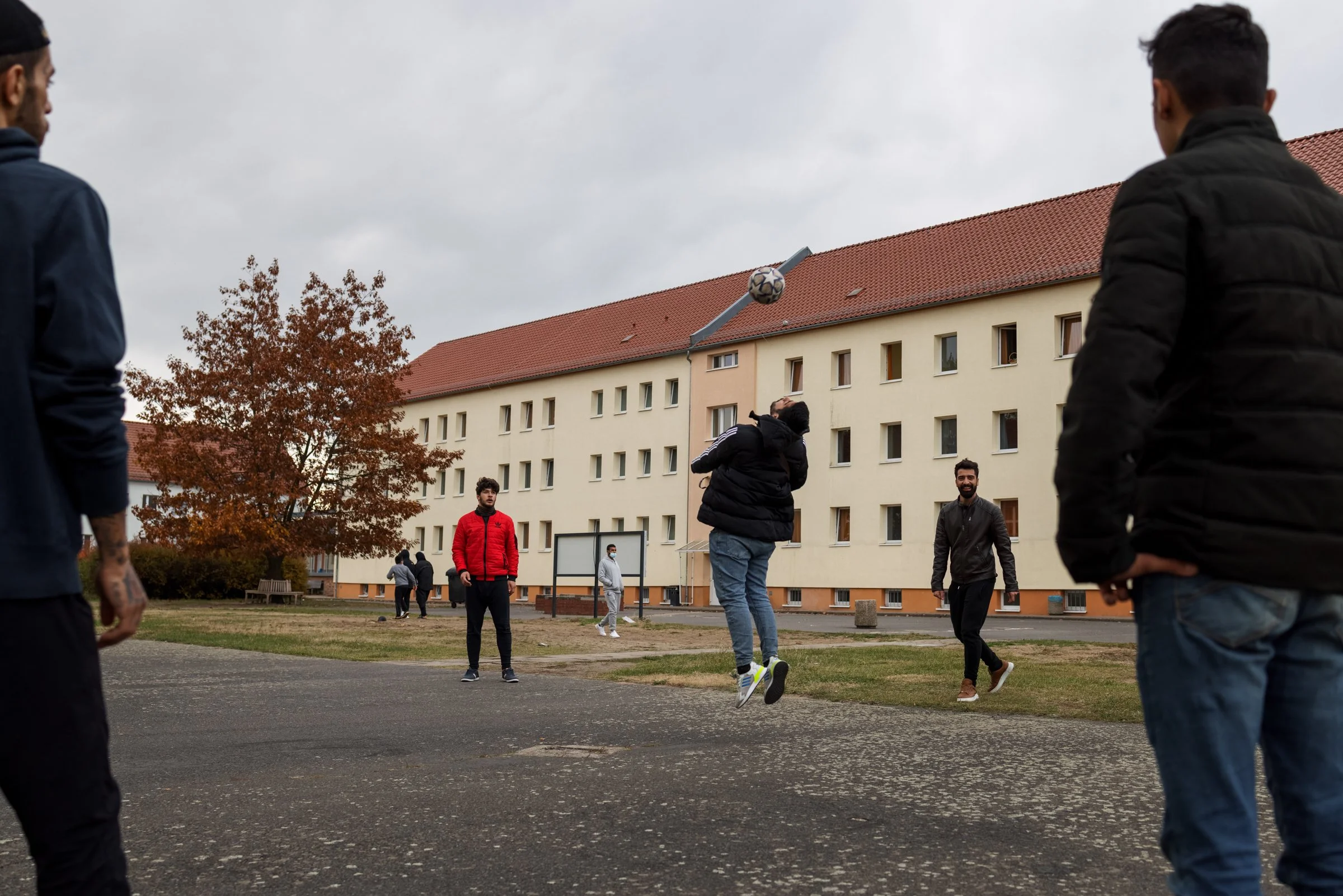 A group of people playing or watching soccer outdoors on a cloudy day in a residential area, with a multi-story building and a tree with autumn-colored leaves in the background.