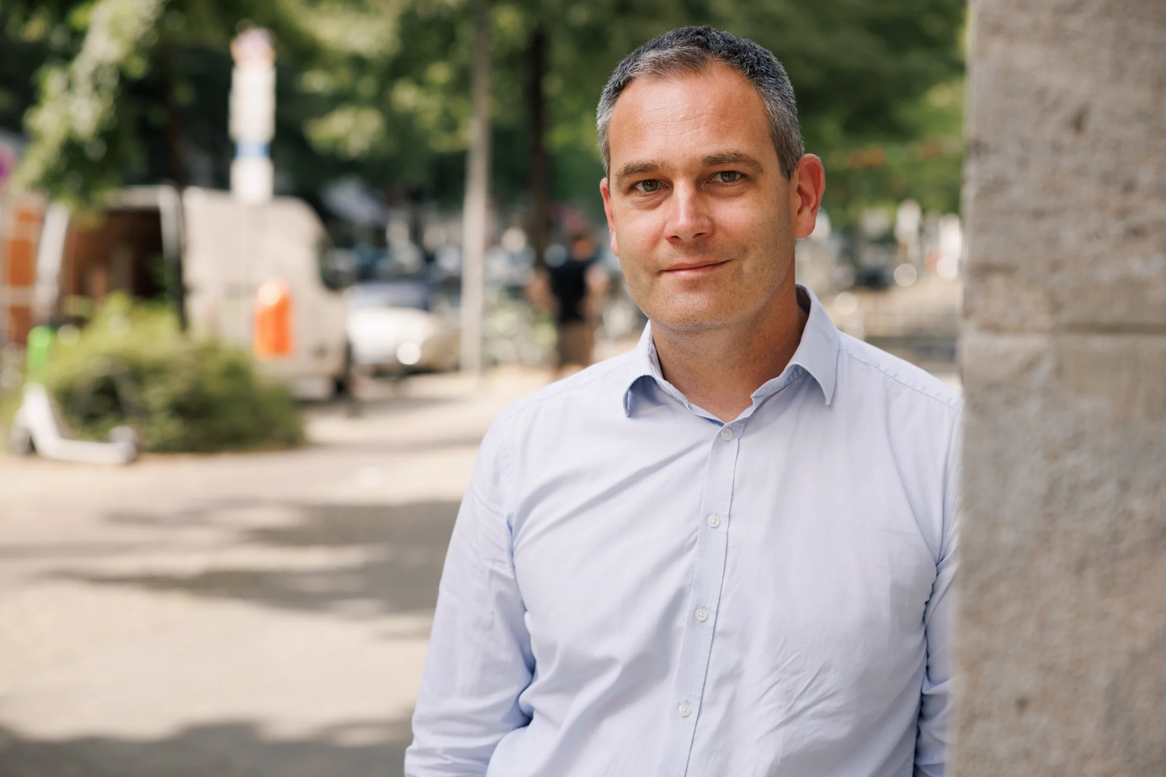 A man with short gray hair wearing a light blue dress shirt, standing outdoors next to a tree with a blurred background of cars and greenery.