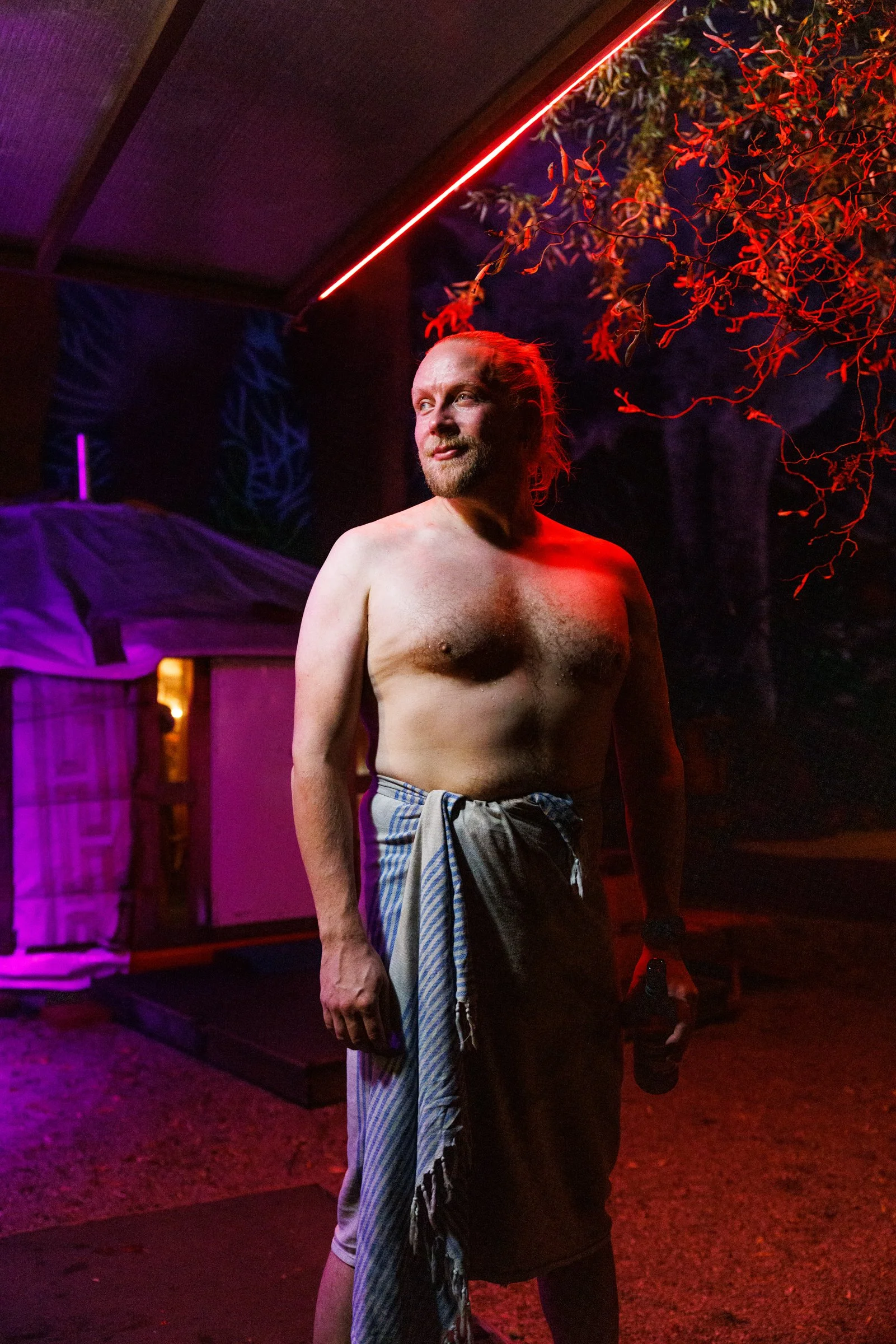 A topless man with a beard and long hair standing outdoors at night, illuminated by red and purple lighting. He is wearing pants with a towel or cloth tied around his waist, with trees and a structure in the background.