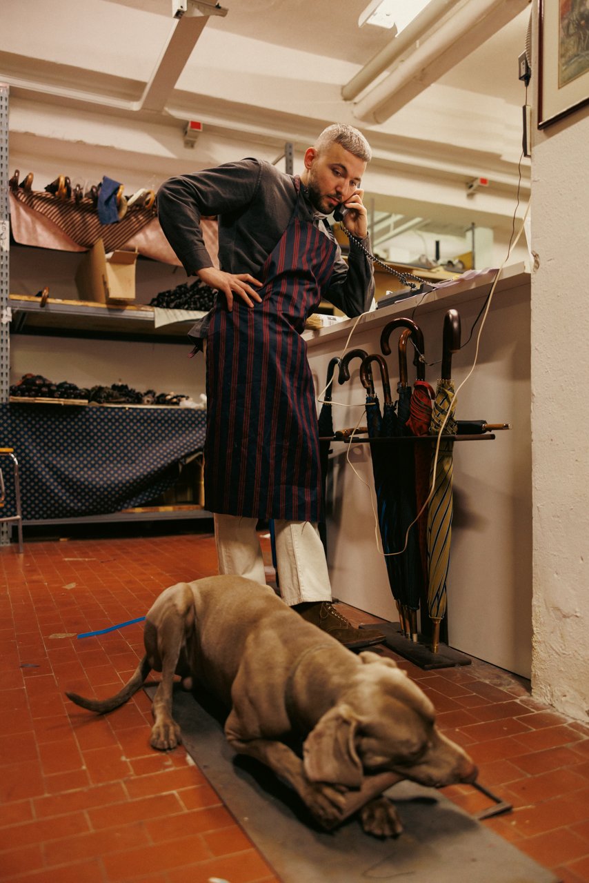 Francesco Maglia, current CEO, wearing a striped apron, standing in the Maglia workshop near finished umbrellas, with a Weimaraner dog nearby.