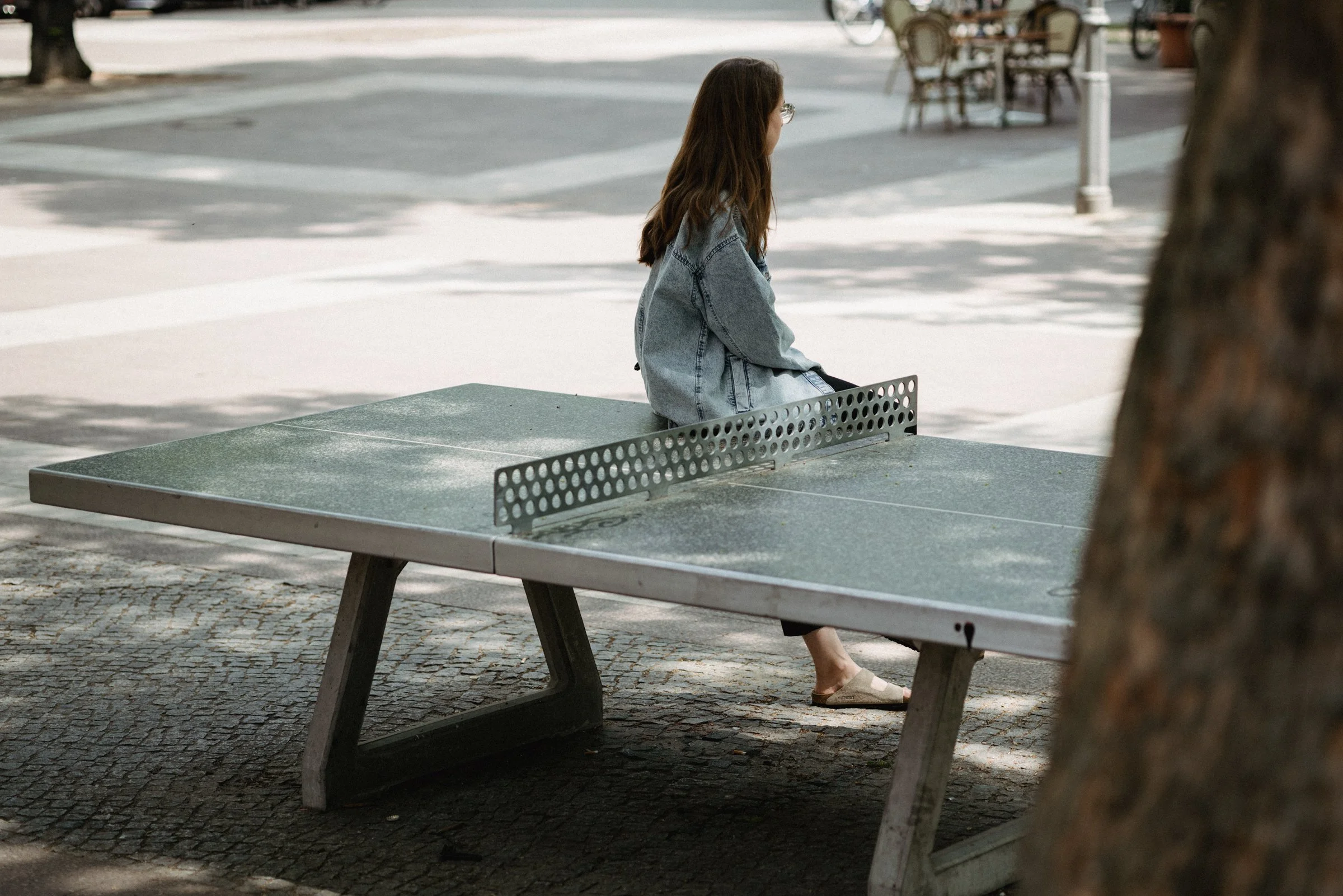 A woman with brown hair and glasses sitting on a park bench, wearing a denim jacket and sandals, in an outdoor urban setting.