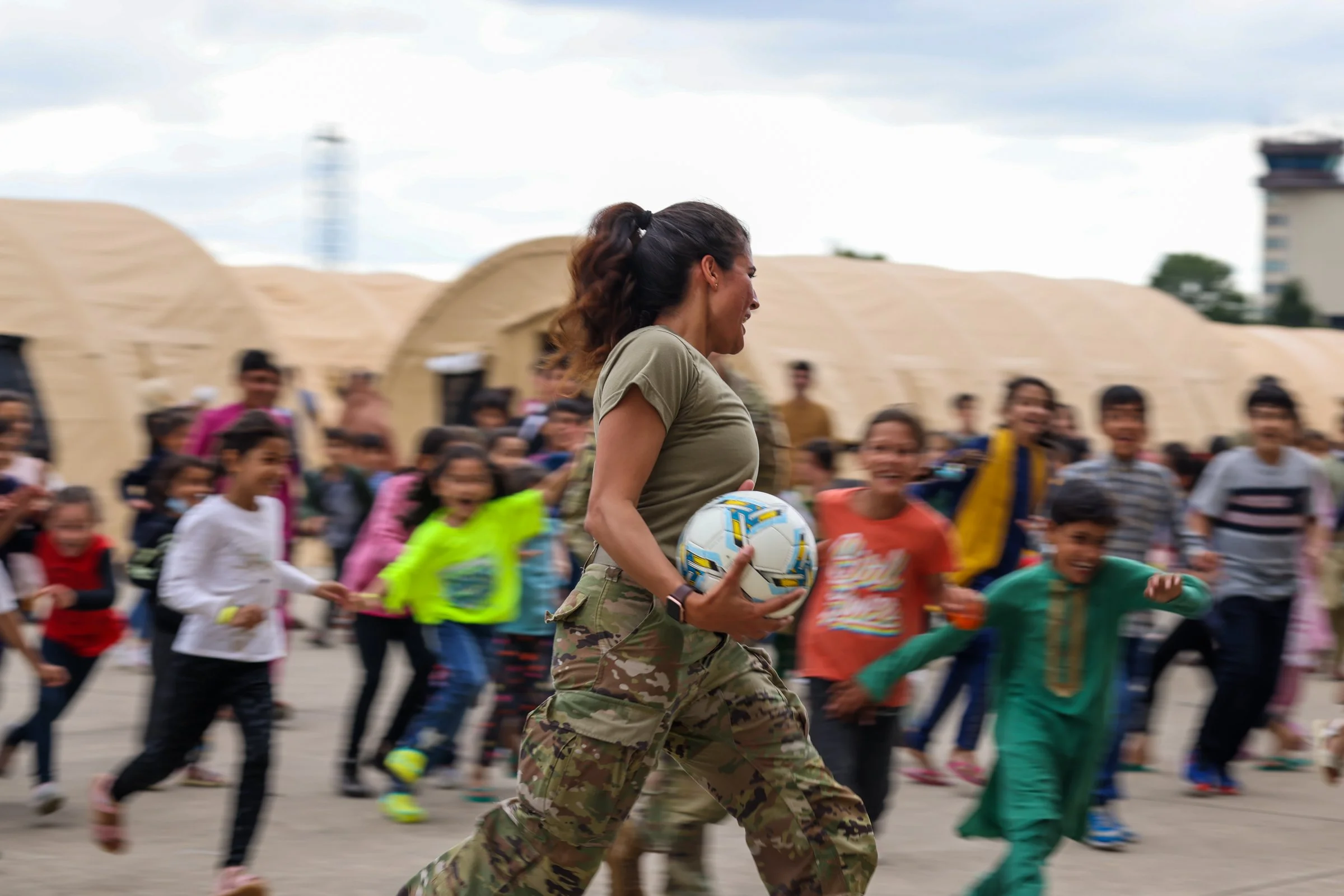 A woman in camouflage military attire running with a group of Afghan children outdoors during daytime, holding a soccer ball  in Ramstein American Airbase in Germany.