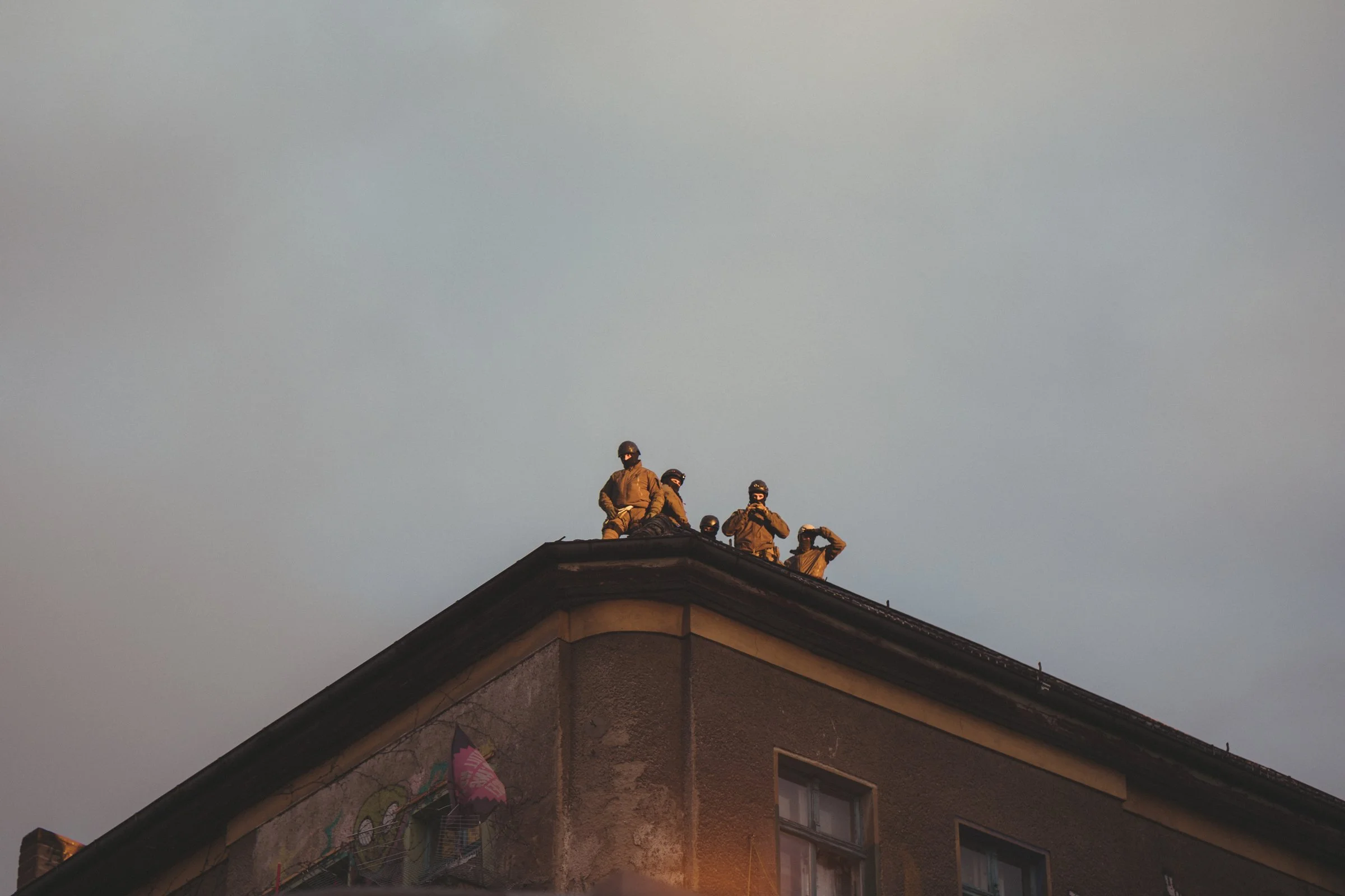 Group of police in full combat gear, standing on the roof of a house, wearing helmets and dark clothing, silhouetted against the evening sky.