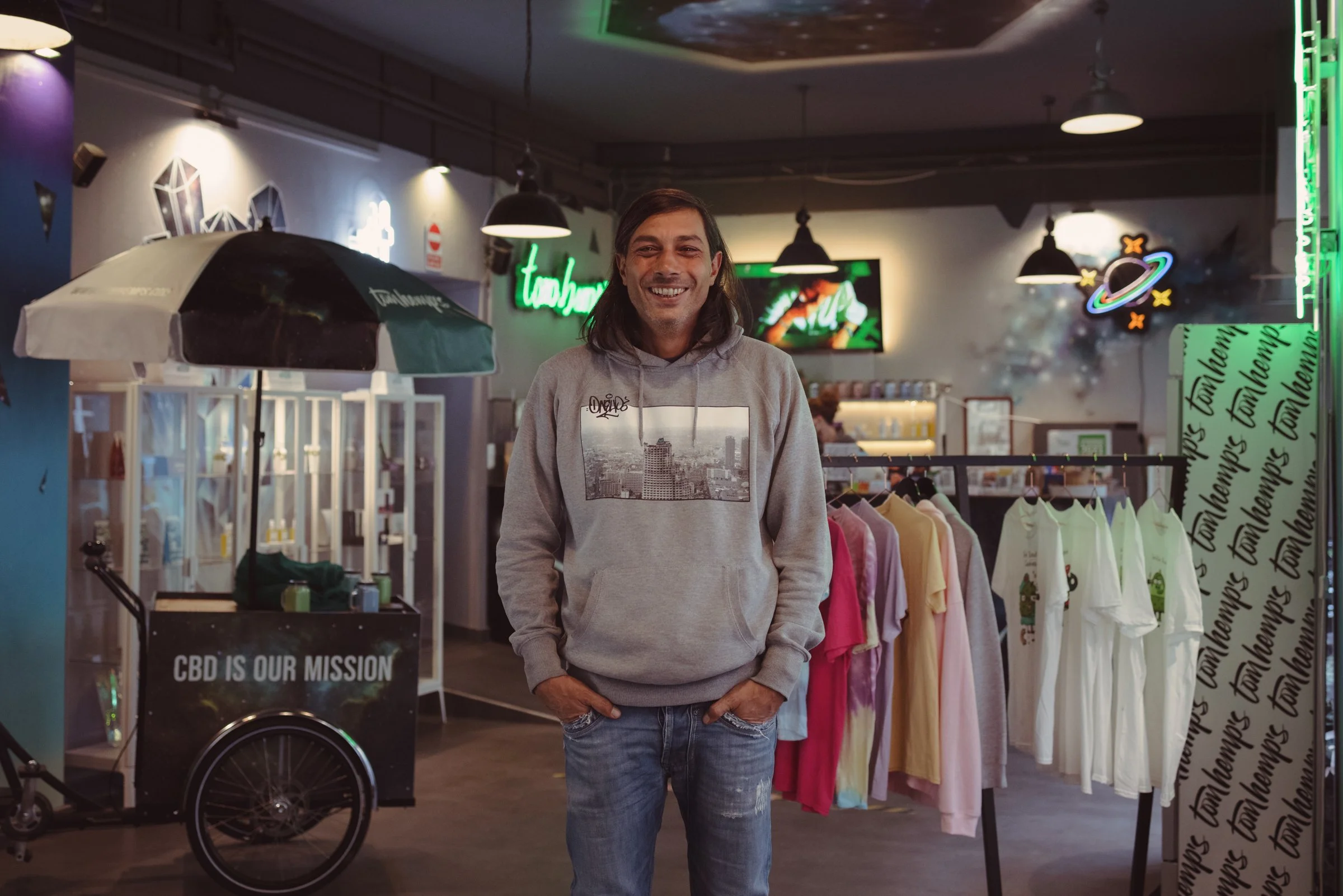 Owner of Tom Hemps in Berlin, smiling, wearing a gray hoodie with a cityscape graphic, standing inside a retail store with colorful clothing racks, neon signs, and space-themed decor.