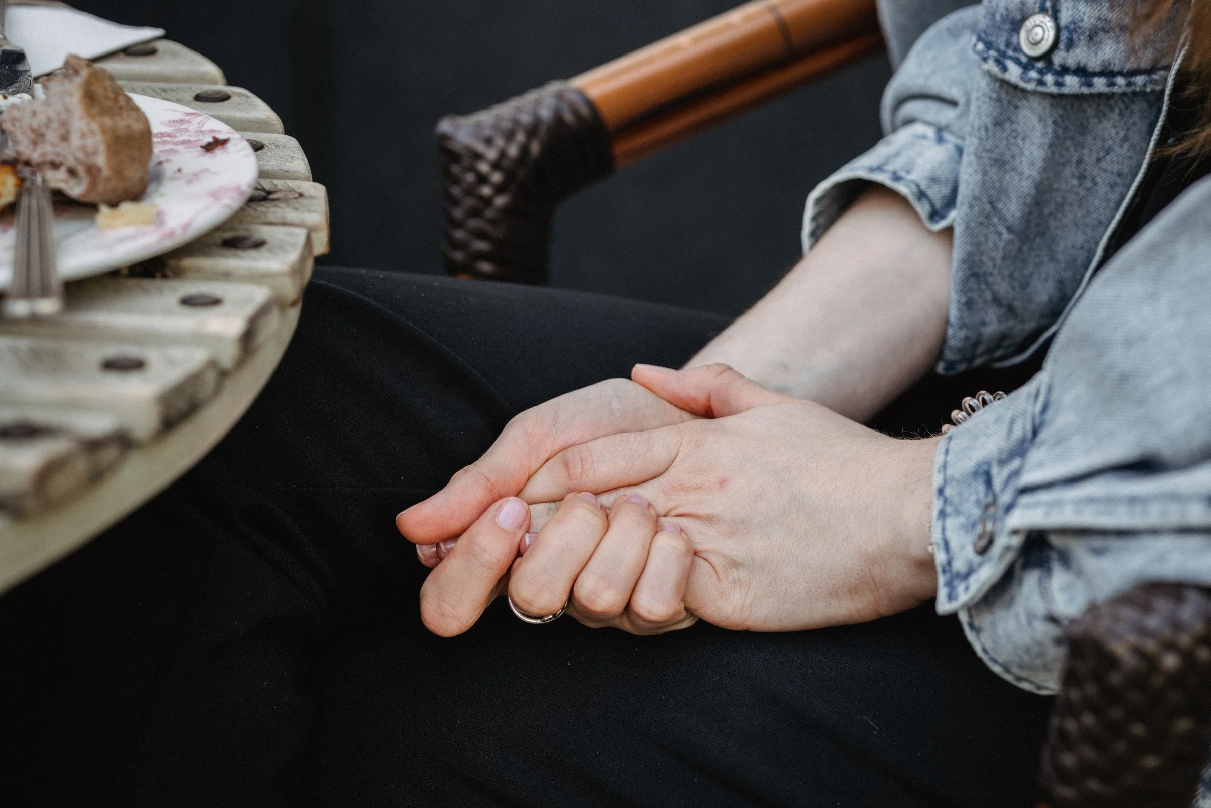Two people holding hands, sitting at a table with a plate of food, in an outdoor setting.