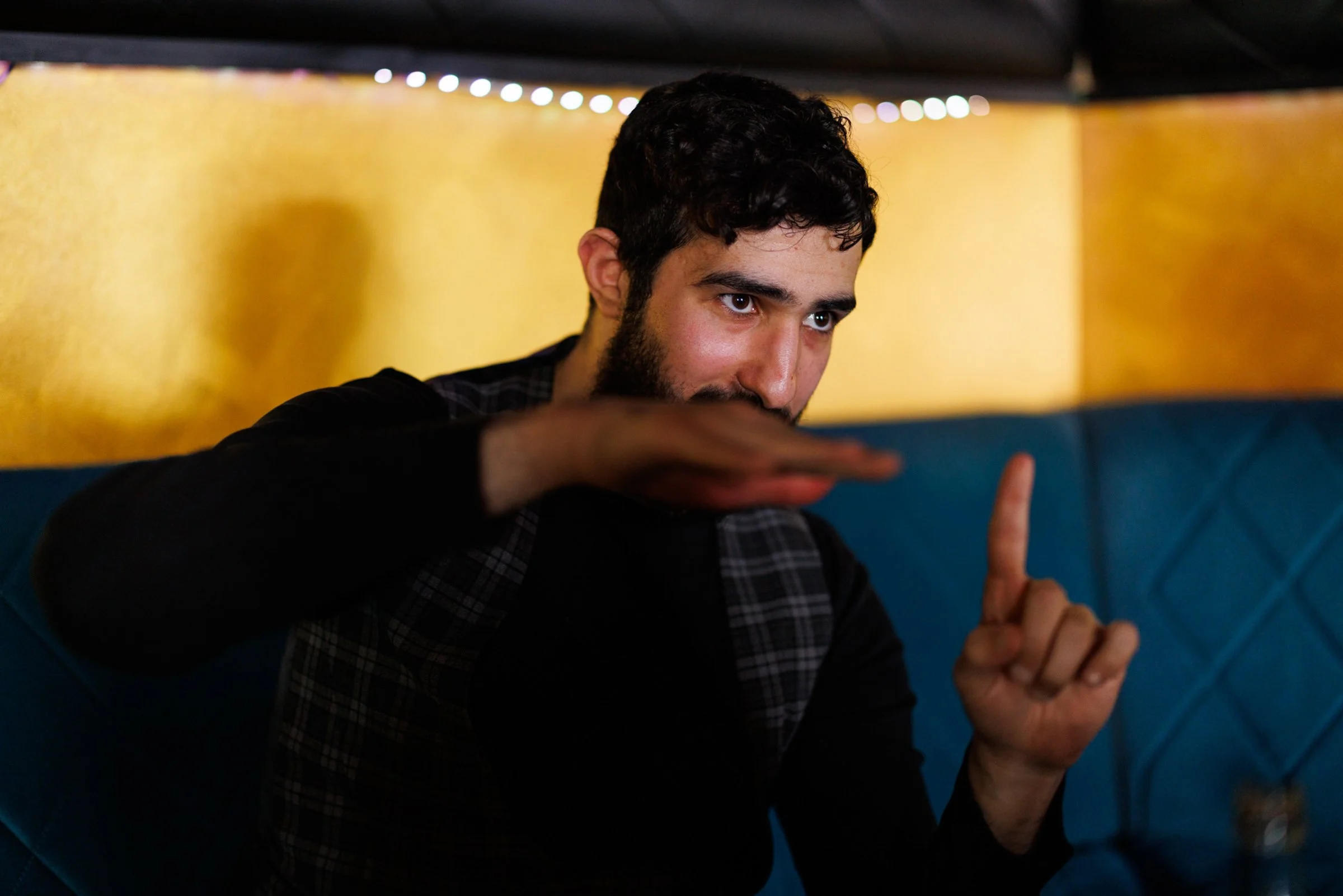 Young man with dark hair and beard, wearing a black shirt and checkered vest, sitting on blue booth with yellow wall behind him, gesturing with his hand.