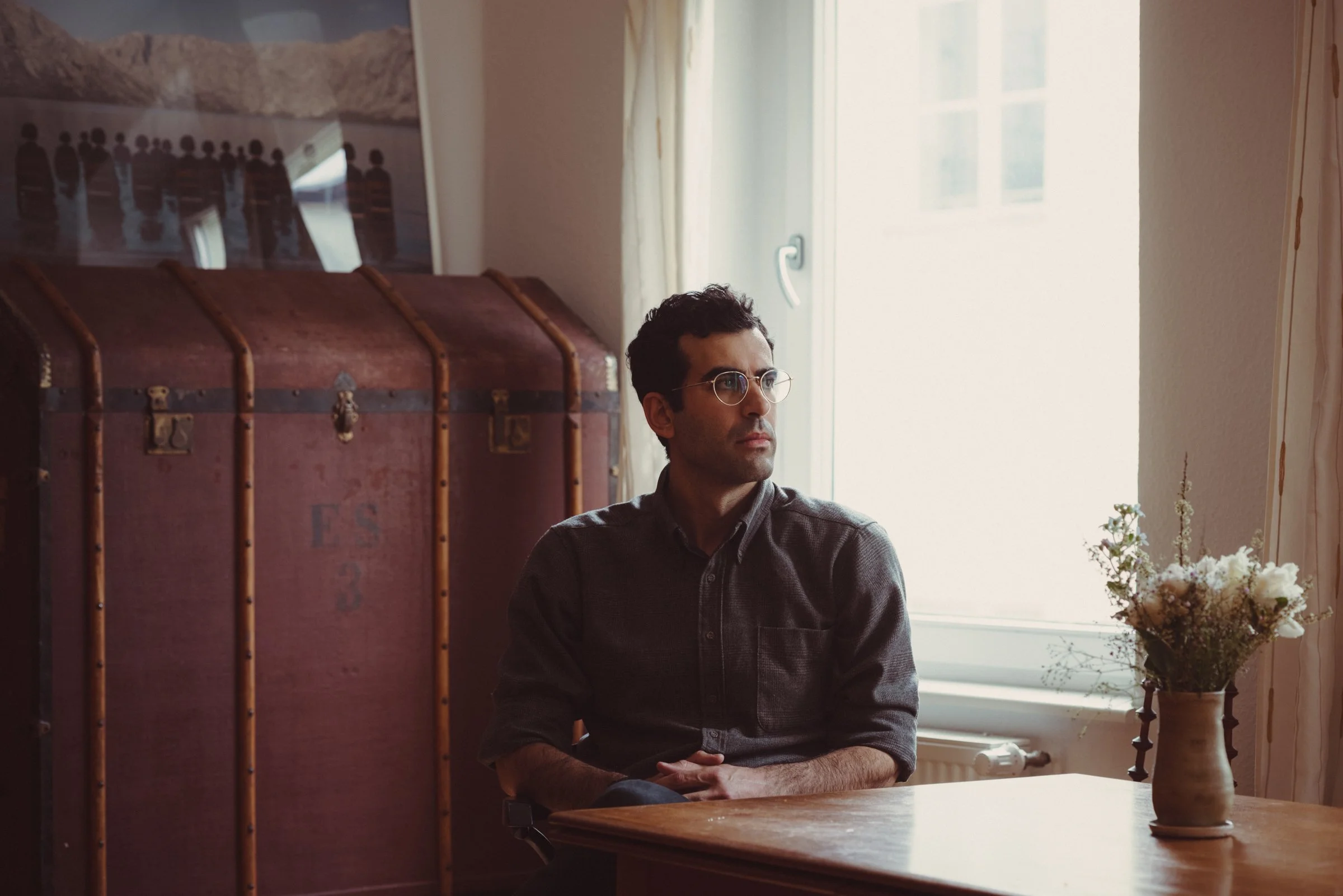 Aaron Benanav, a man with dark hair, glasses, and a gray shirt sitting at a wooden table near a window with white curtains, a large chest behind him, and a flower arrangement in a vase on the table.