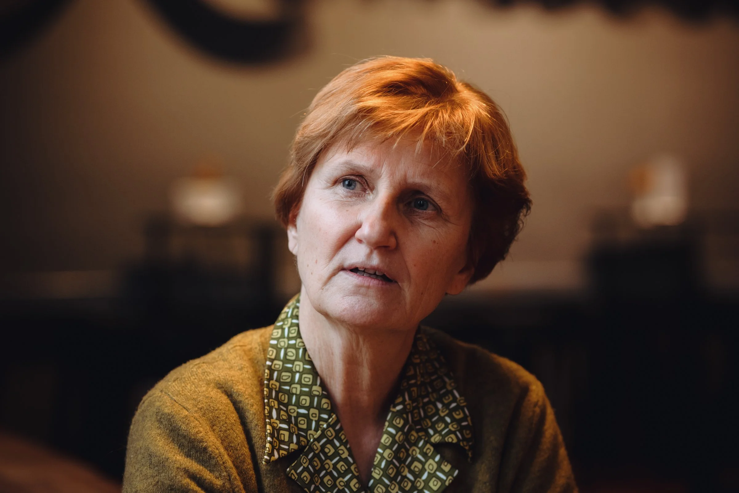 A woman with short reddish hair and blue eyes, wearing a patterned shirt and a brown sweater, is looking to her left in an indoor setting with blurred background.