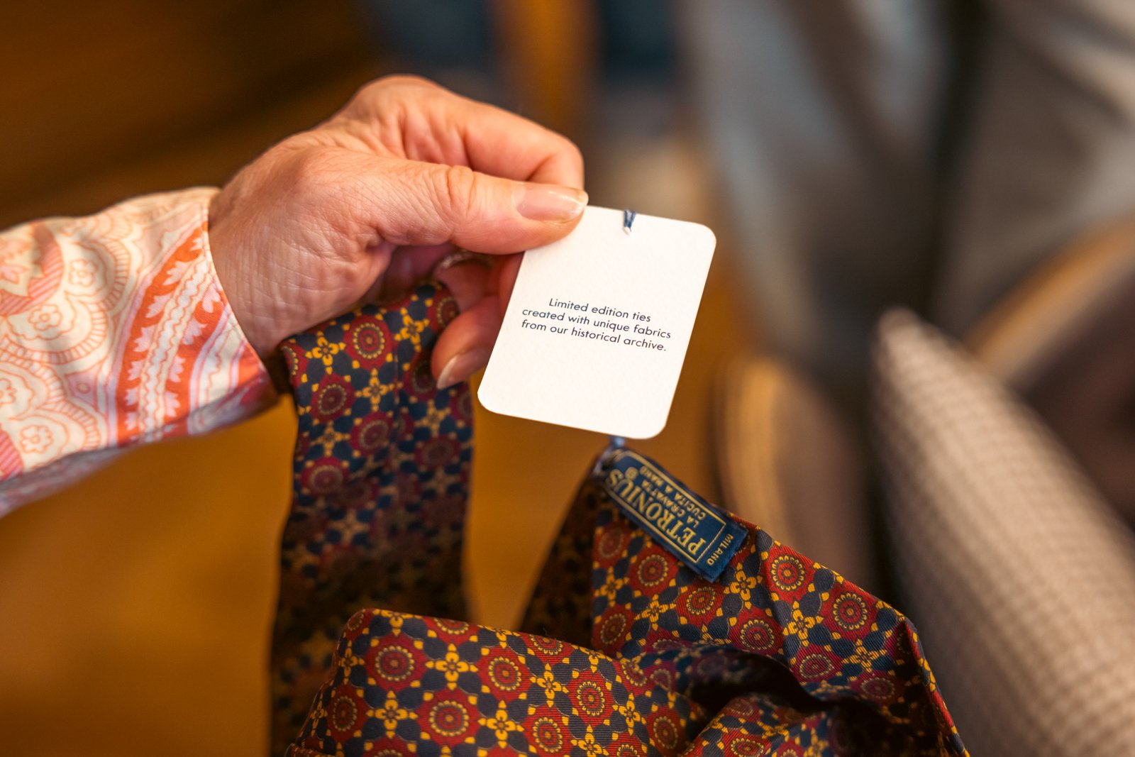 Close-up of a hand holding the label attached to a richly patterned Petronius 1926 silk tie.