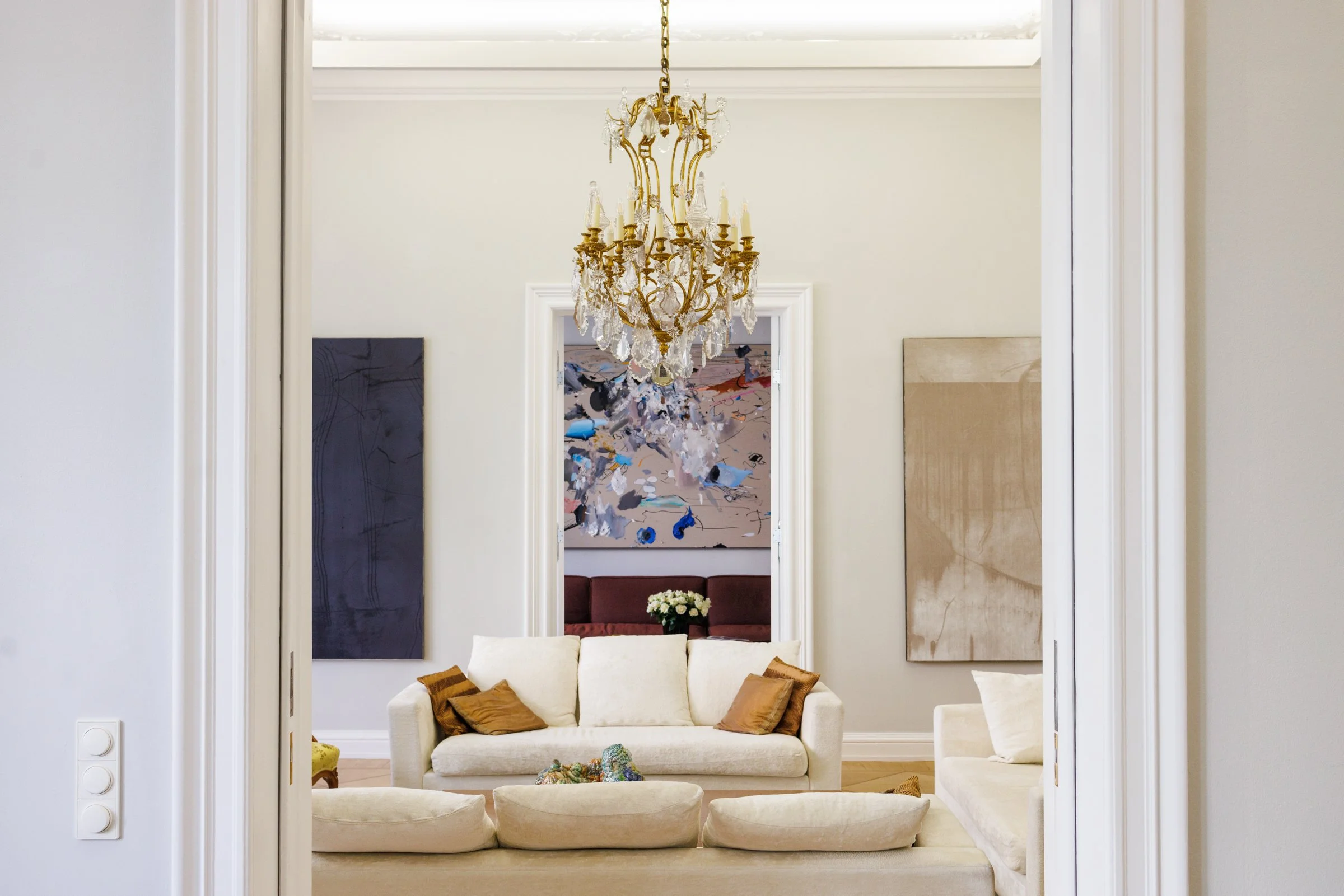Living room with white sofa and ottomans, gold chandelier, abstract artwork, and a vase of white flowers, viewed through doorway.