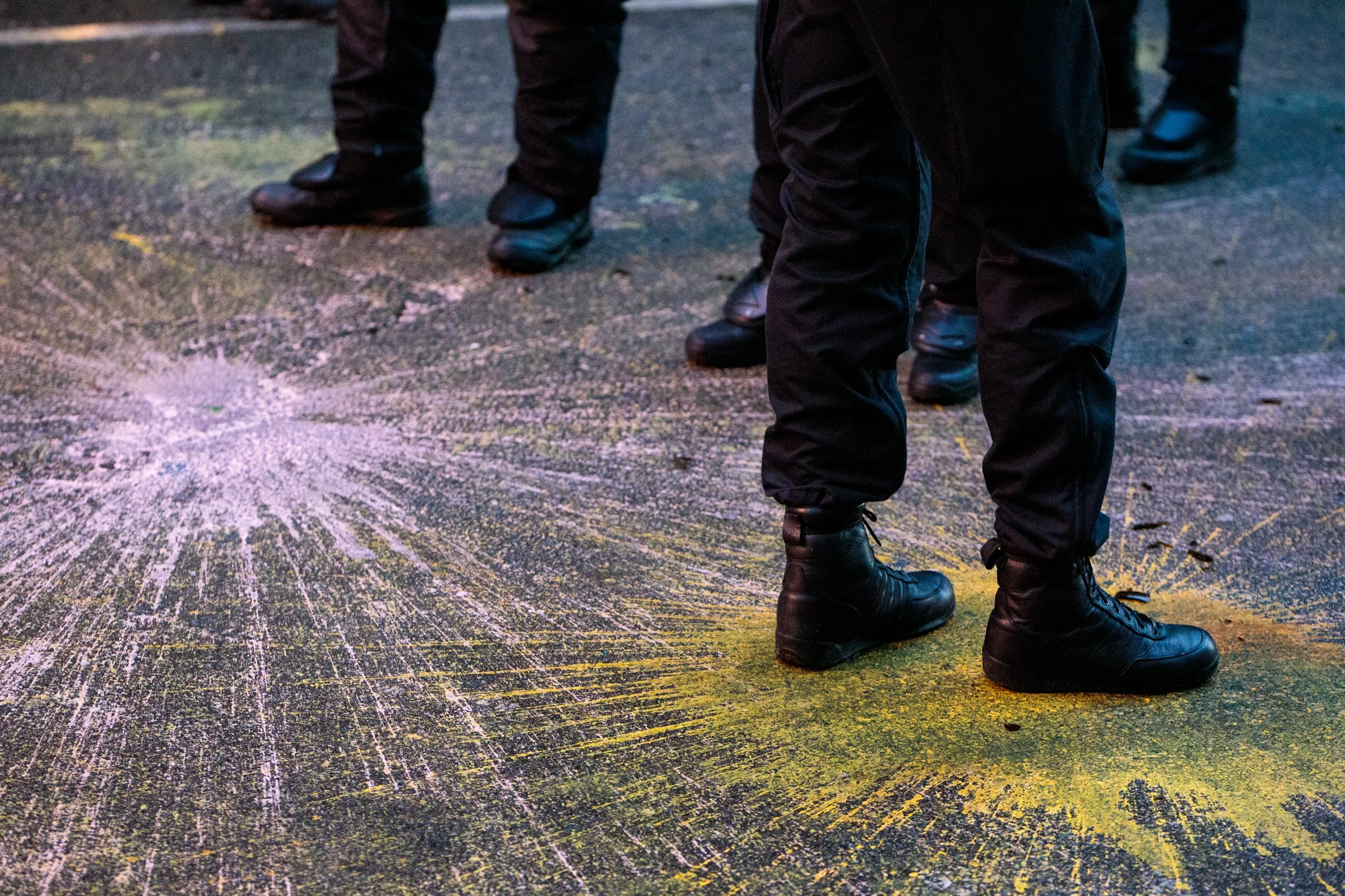 Close-up of police officers in black uniforms and boots standing on a street with yellow and pink chalk markings.