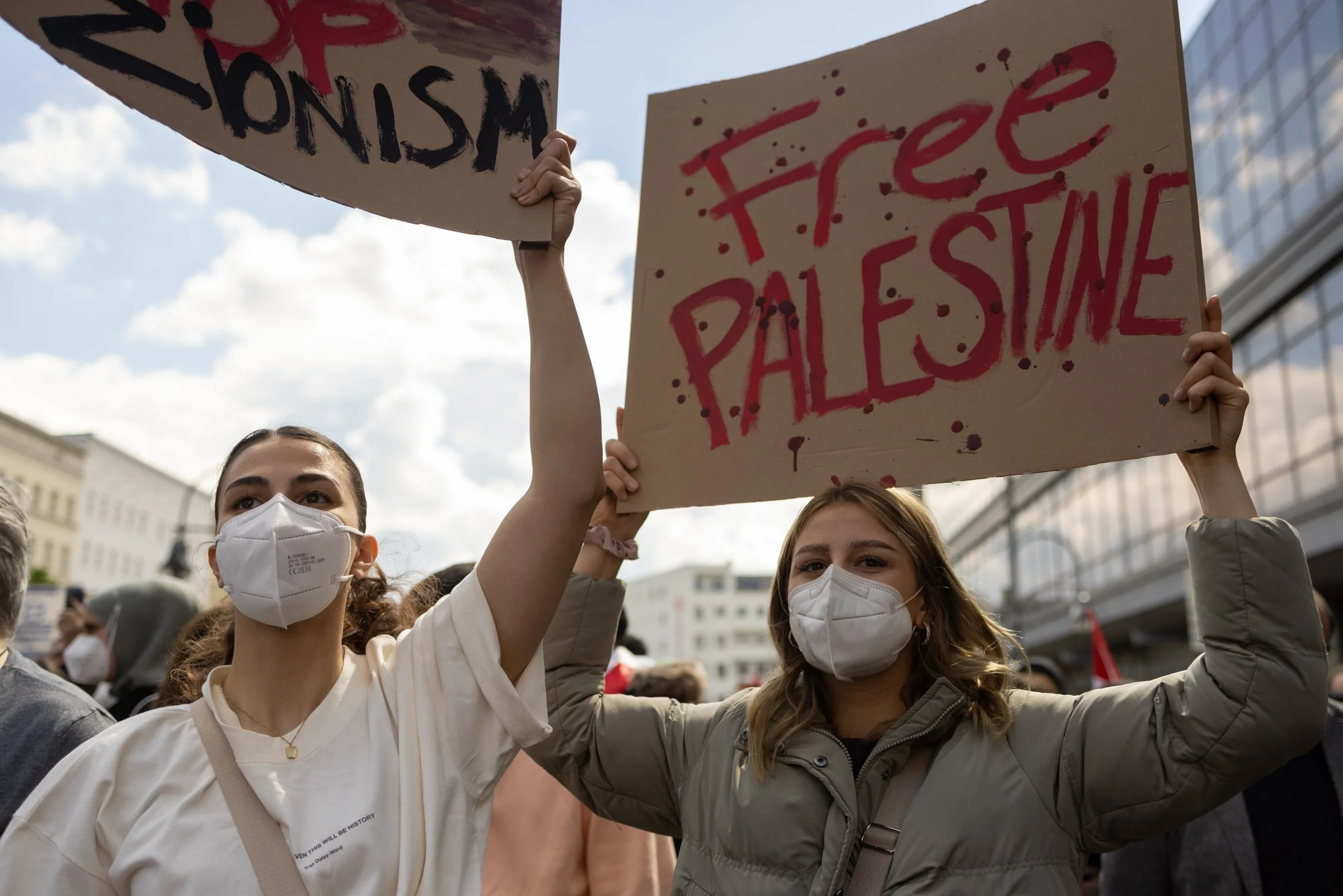 Two young women wearing face masks at a protest, holding handmade signs in the air. One sign says 'Justice' and the other says 'Palocene' with red paint splatters. The women are standing in a crowd outdoors with a building visible in the background.