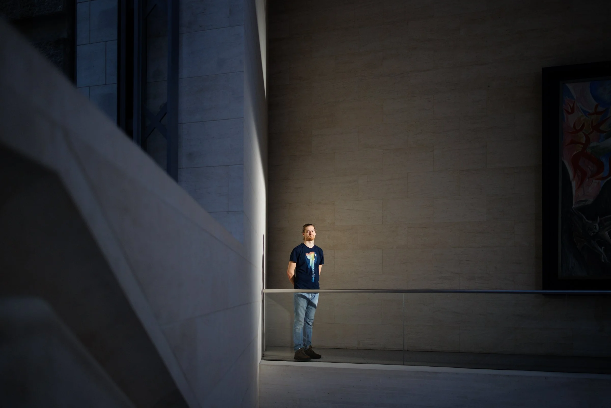 A man with a beard wearing a black t-shirt with a colorful graphic stands in front of a large mirror in a modern, minimalistic building with light-colored walls and a dark ceiling.