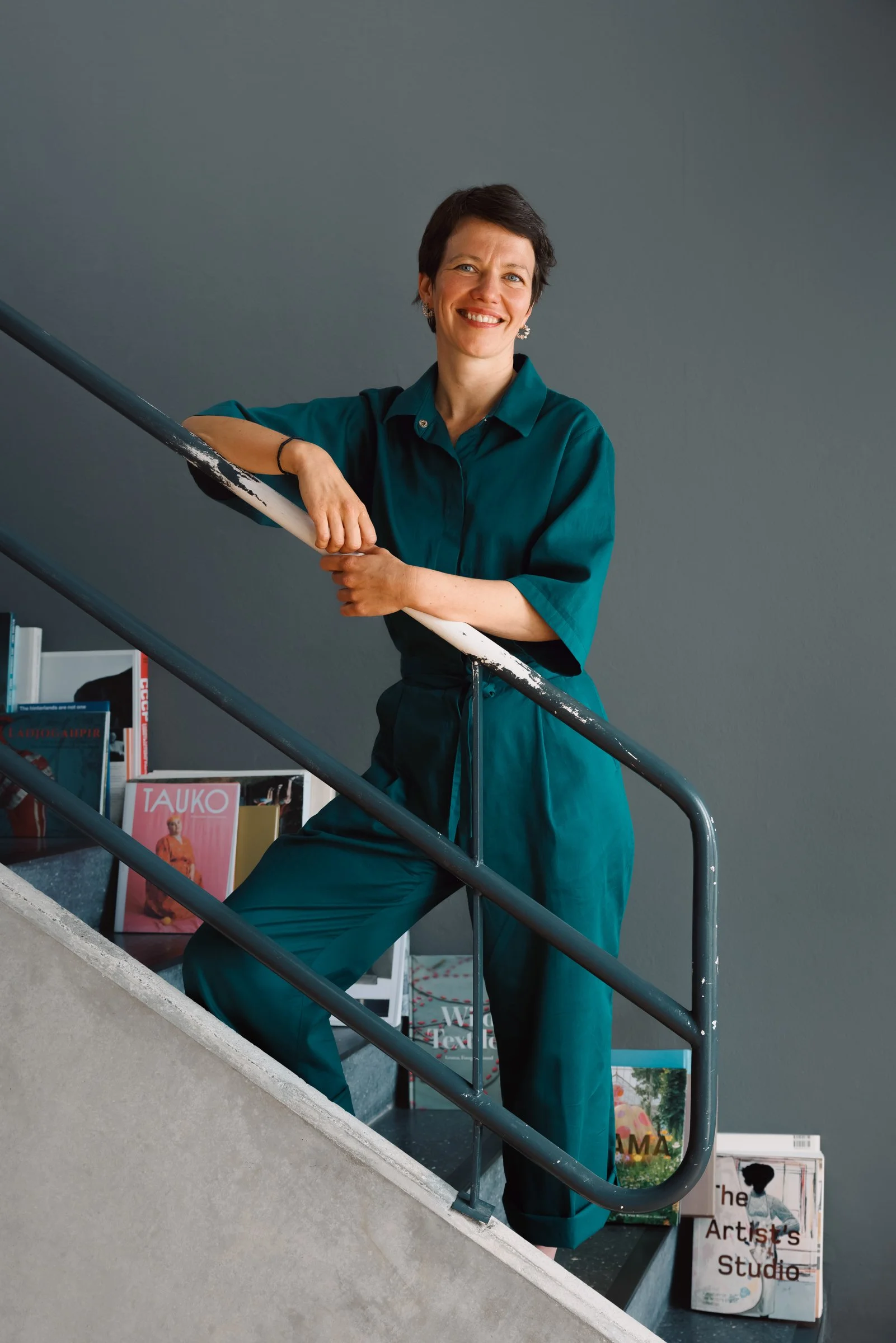 A person with short dark hair and wearing teal clothing is sitting on a staircase railing and smiling at the camera. Behind them is a gray wall with a bookshelf and magazines, including one titled 'The Artist's Studio'.