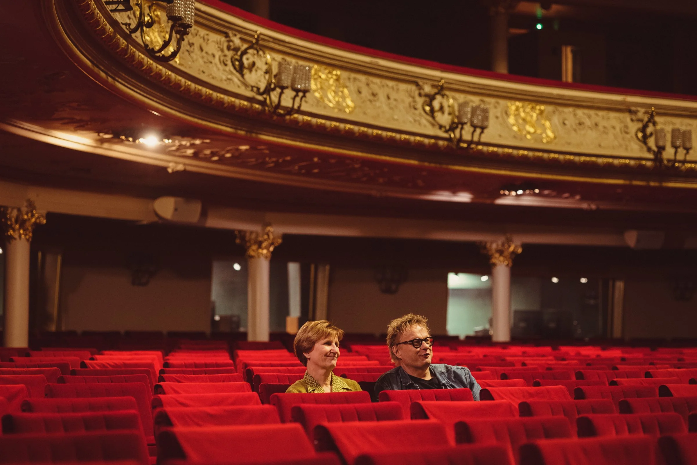 Two women sitting in a theater with red seats, ornate gold and cream balcony above, and two people in the foreground.
