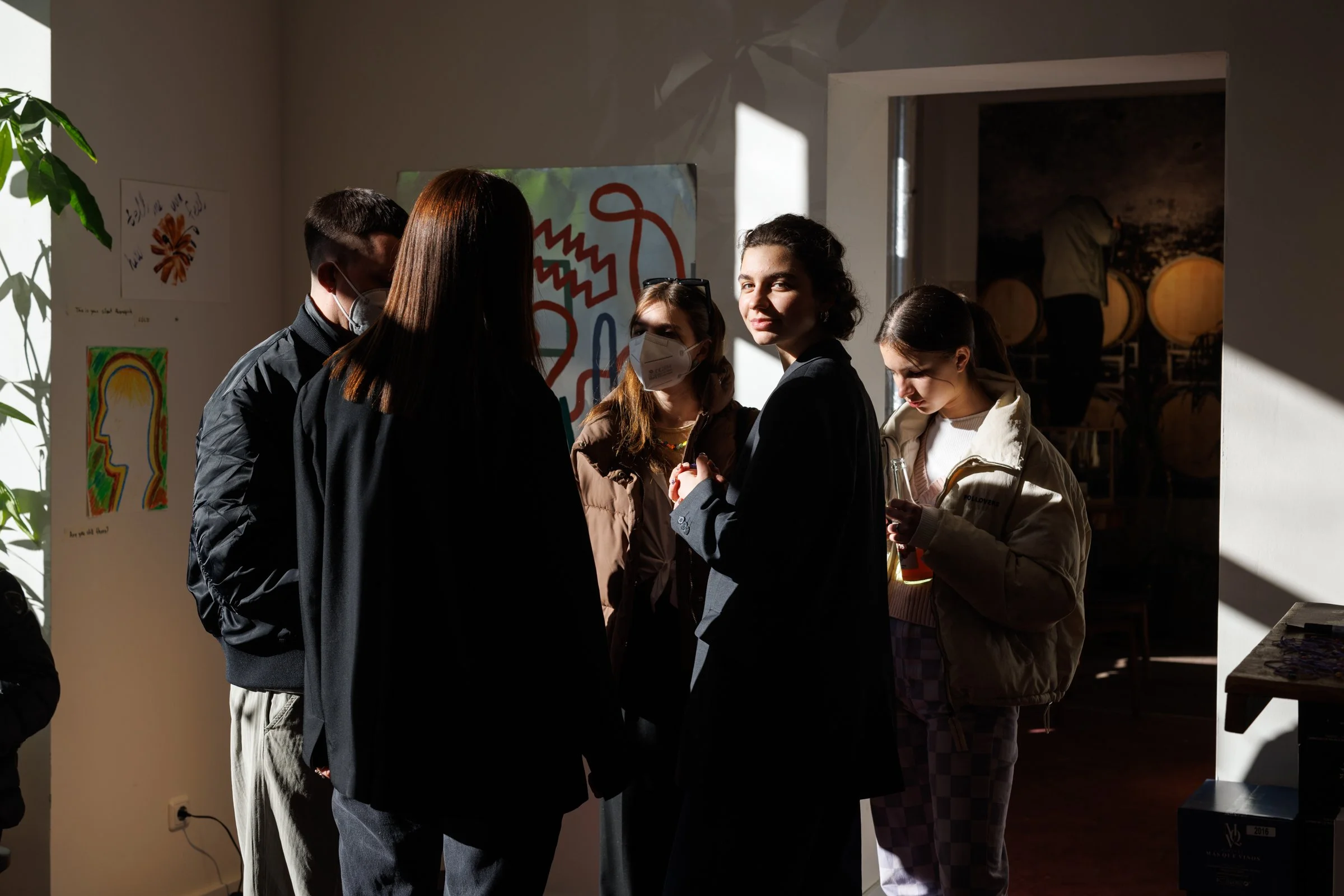 Group of people standing in a room, some wearing face masks, engaging in conversation with sunlight casting shadows. Artwork and wine barrels are visible in the background.