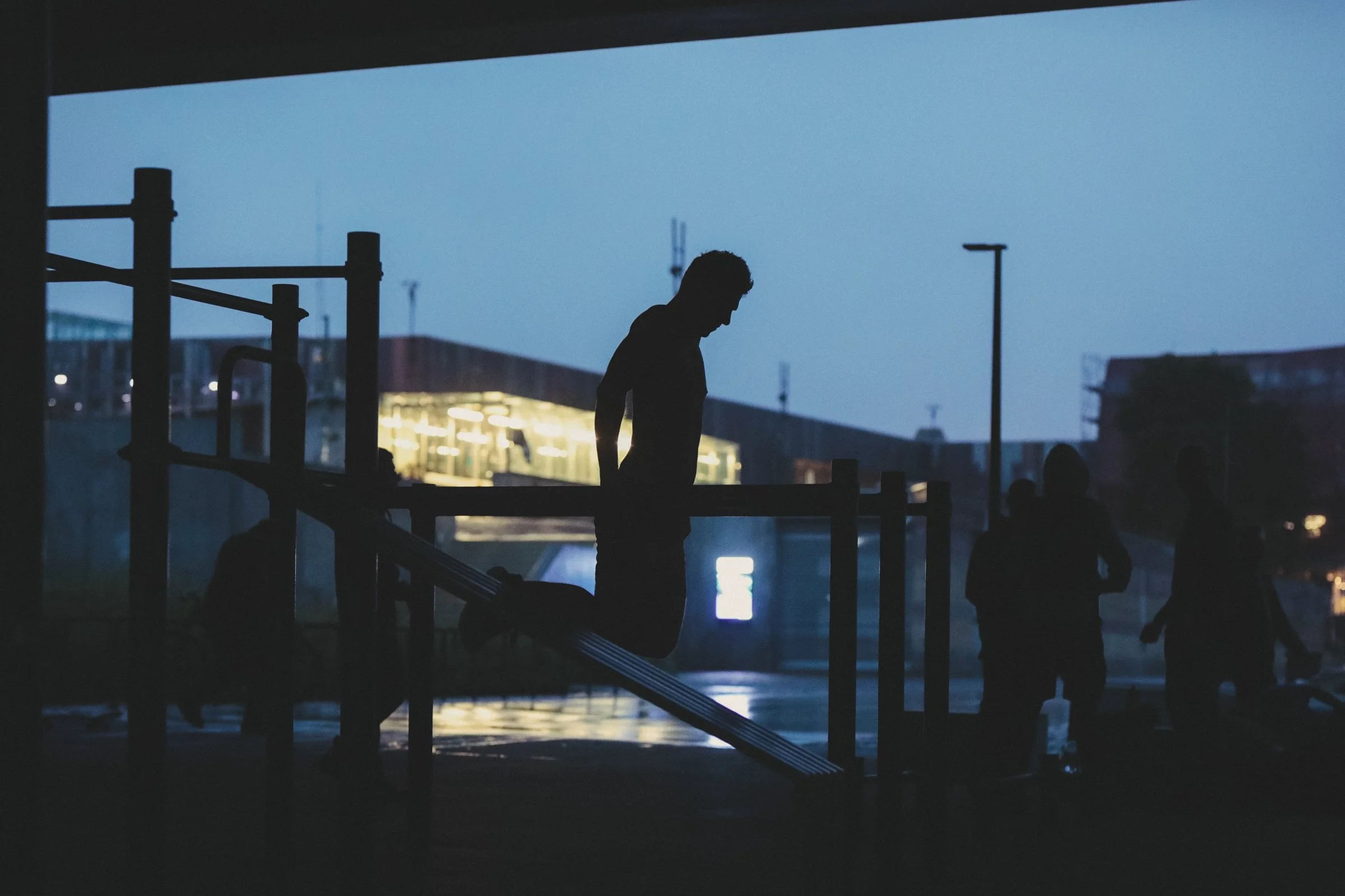 Silhouetted man excercising near a body of water during twilight or early evening, with bridges, buildings, and streetlights in the background.