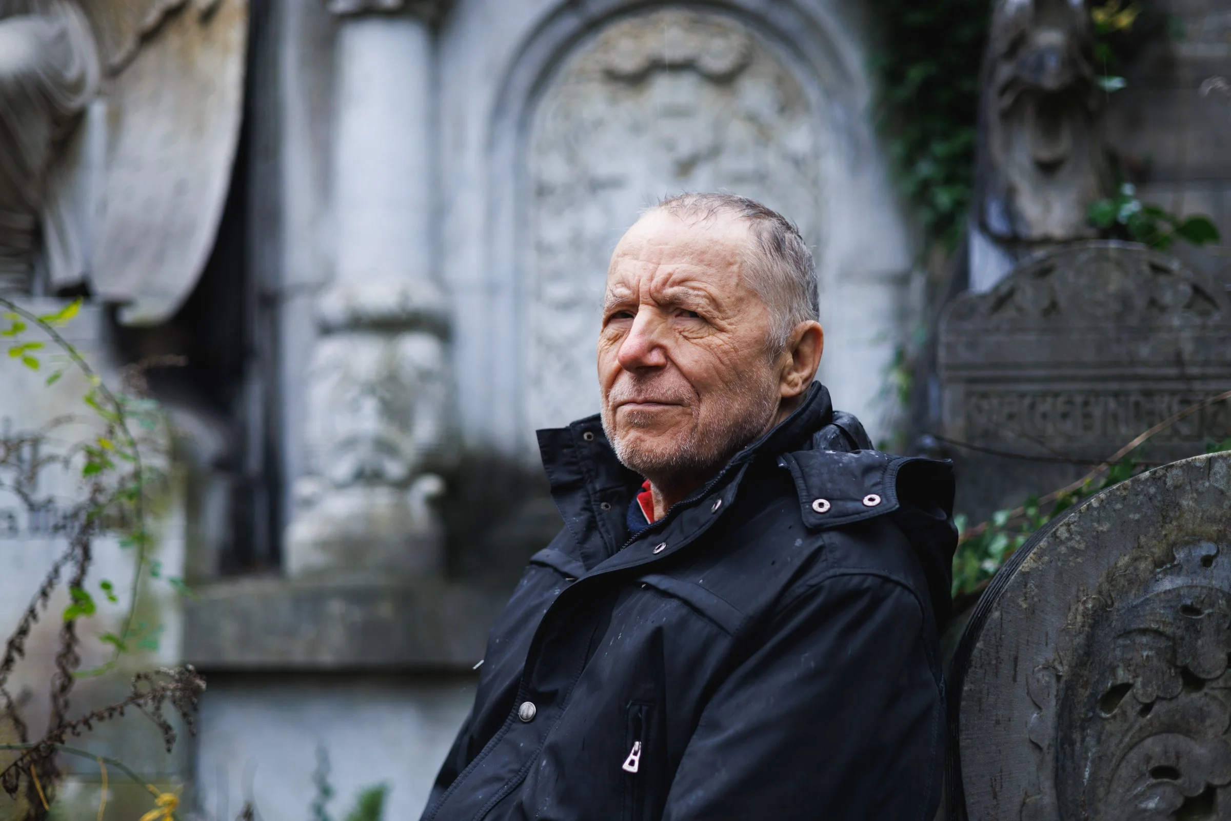 An elderly man with short gray hair and a beard sitting outdoors by a stone wall, wearing a black jacket, with a blurred stone archway and greenery in the background.