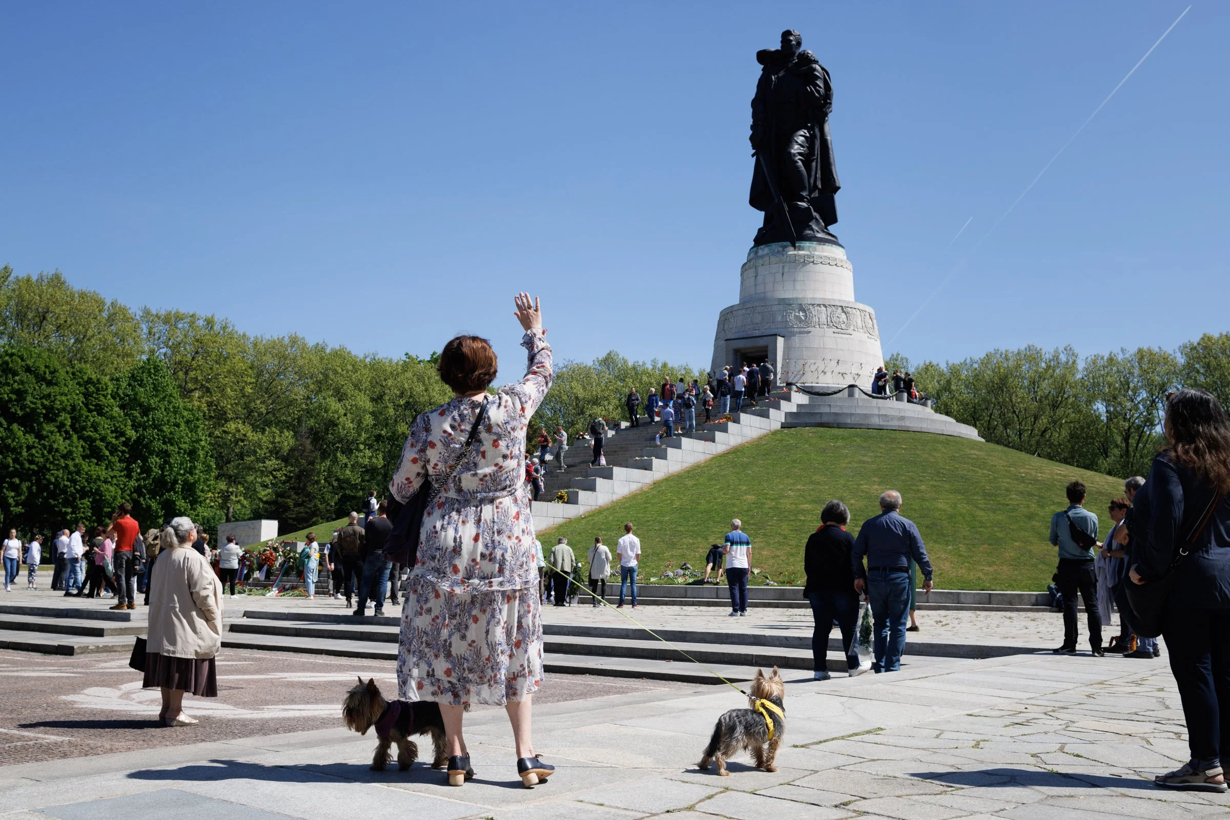 People gathered around the Tsar Cannon at the Kremlin, with a woman in a floral dress waving, others standing and walking on the stairs, and a large statue in the background under a clear blue sky.
