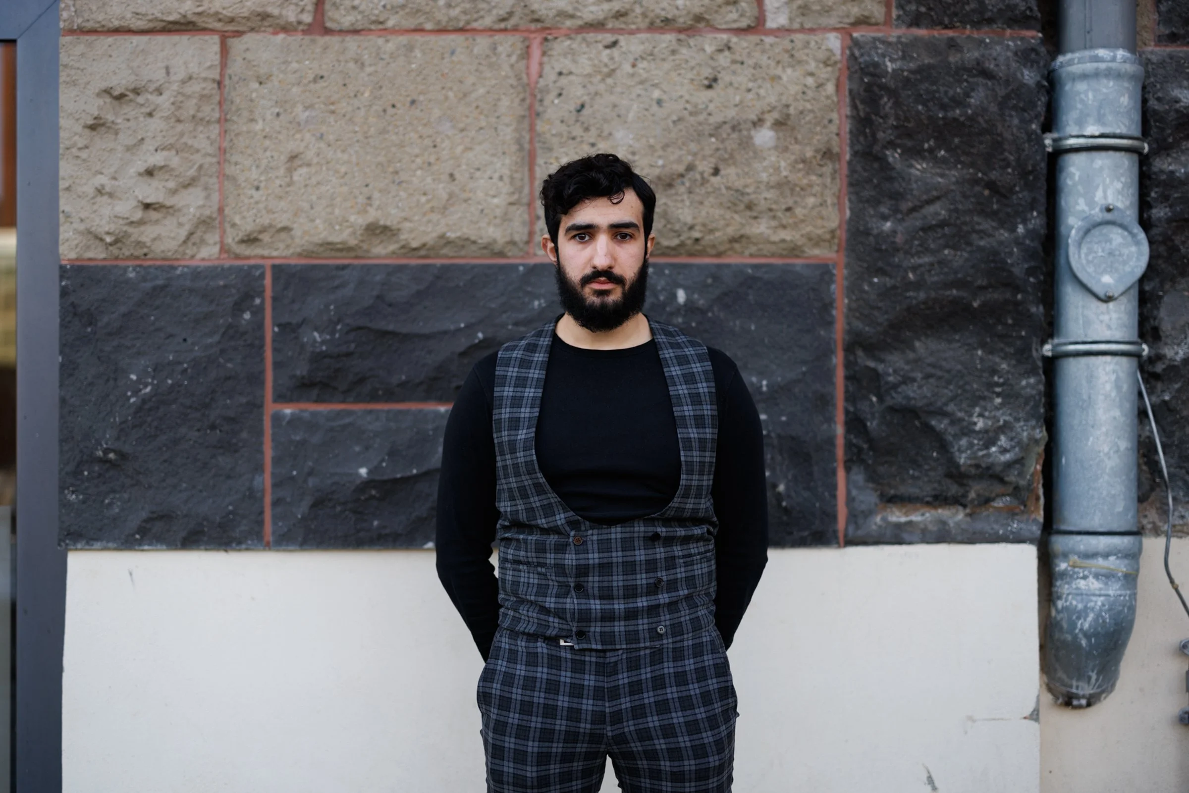 A young man with dark hair and a beard standing in front of a stone wall with a pipe to the right, wearing a black long-sleeve shirt and plaid pants with suspenders.