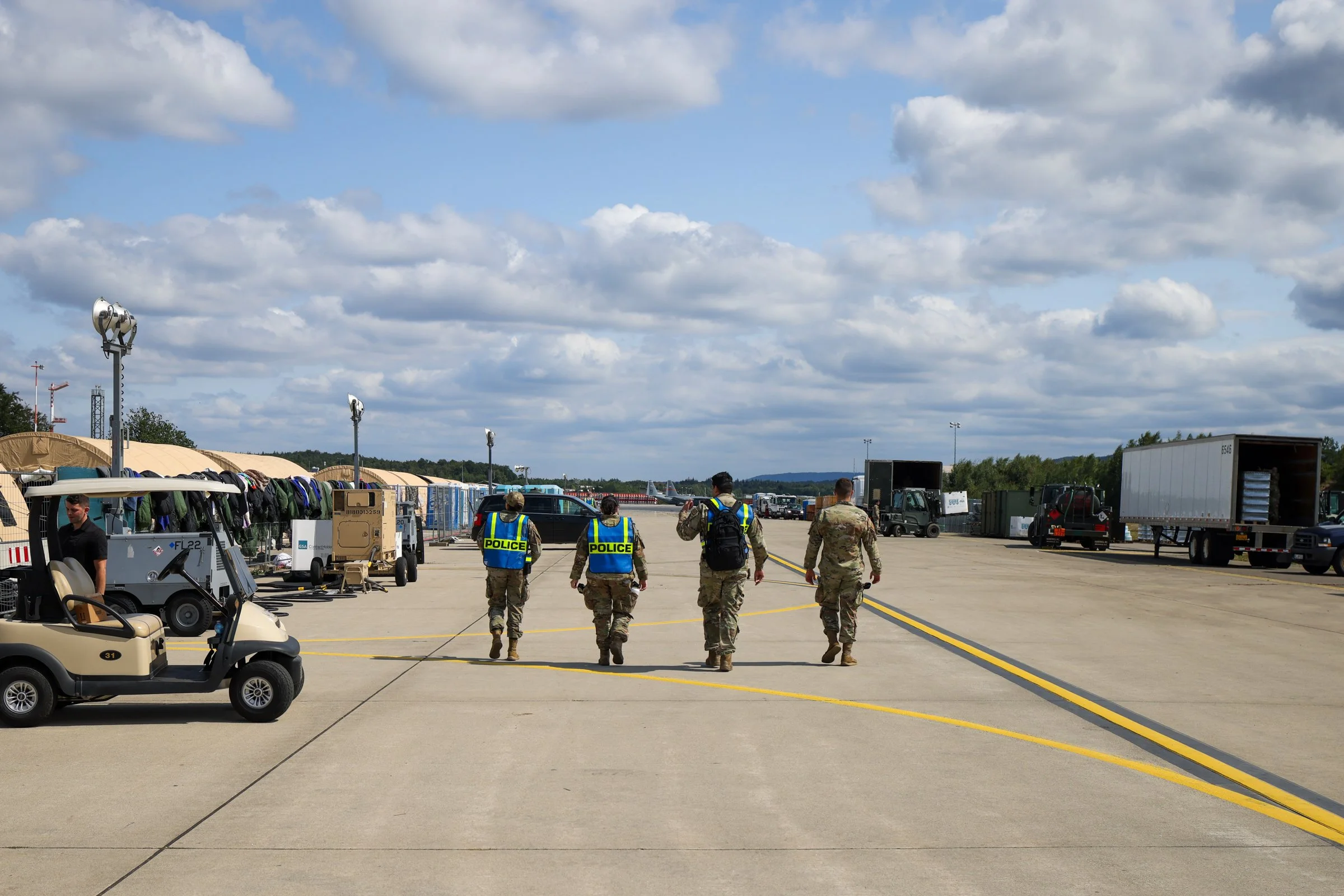 Military personnel walking on an airfield, with various vehicles and equipment around, under partly cloudy skies in Ramstein American Airbase in Germany.