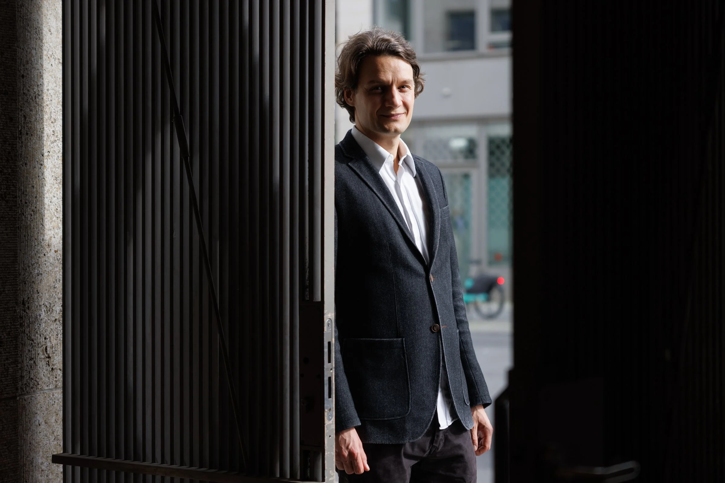 A man in a dark blazer and white shirt standing by a doorway, smiling slightly, with an urban street scene in the background.