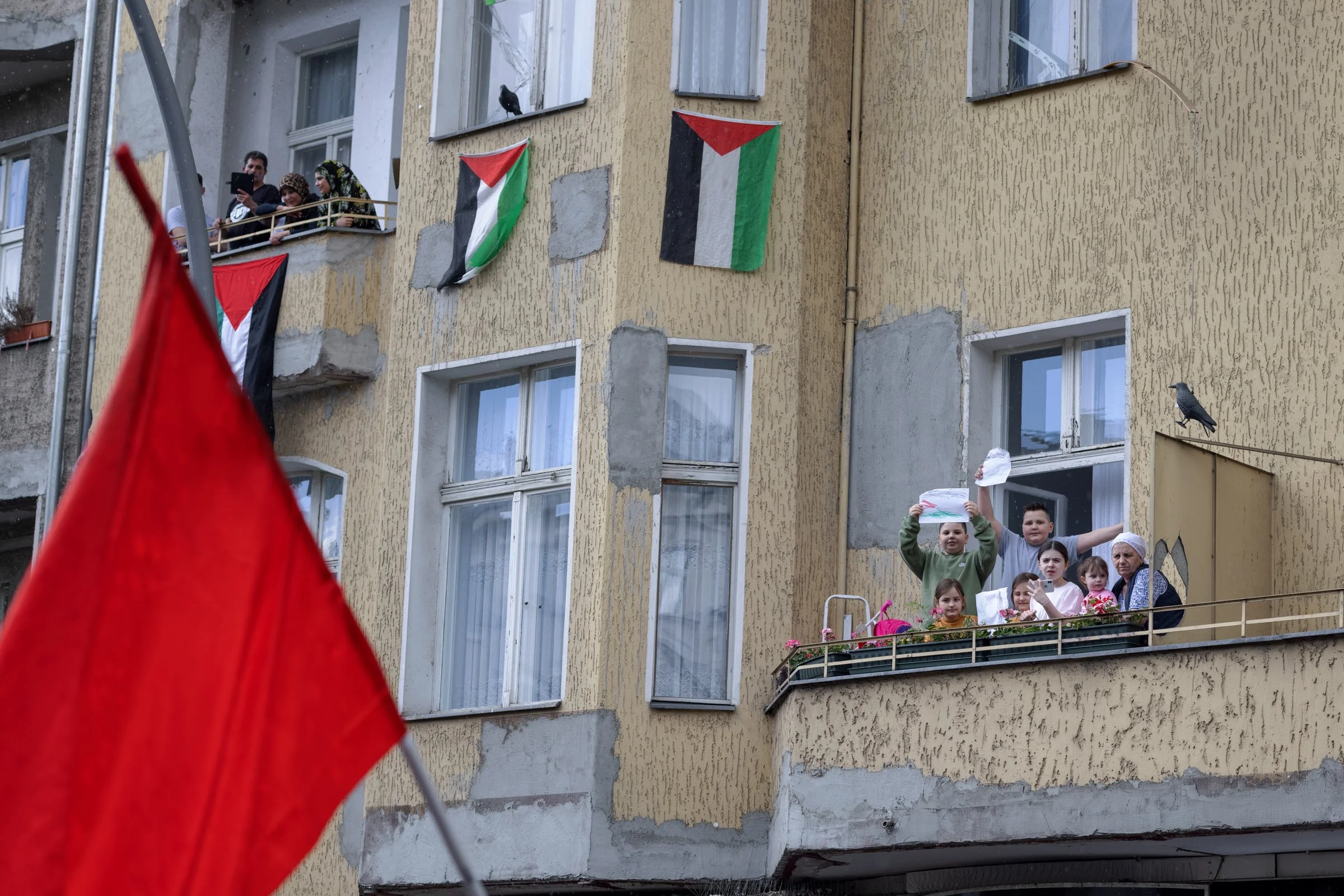People on a balcony decorated with Palestinian flags, some holding signs and children standing in front of the building, with a red flag in the foreground.